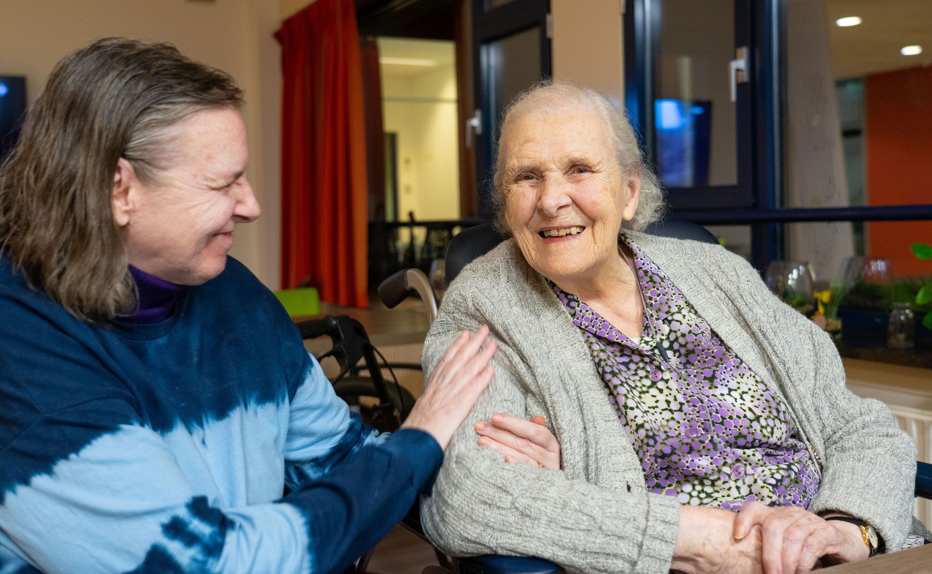 Older woman smiles as a caregiver places a hand on her shoulder indoors.
