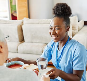 Woman in ponytail talks to an older woman with gray hair on a couch indoors; smiles, light.