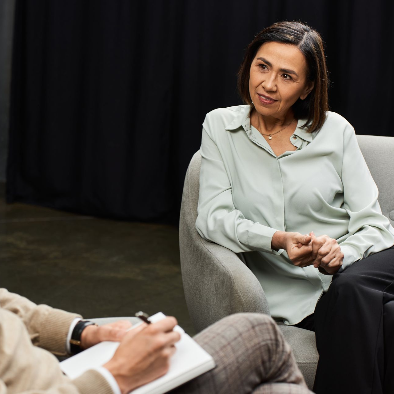 A person sits on a couch as another person takes notes on a clipboard.