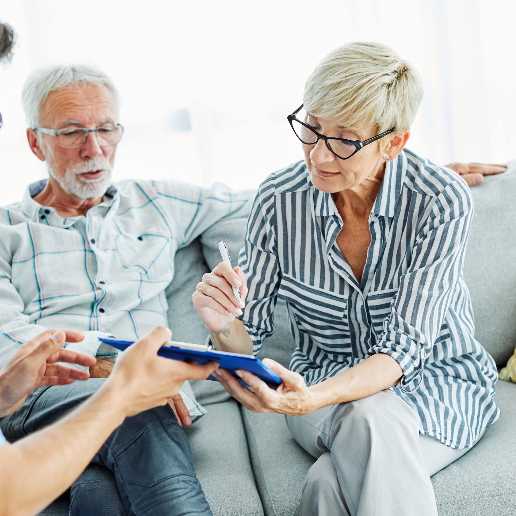 Blonde therapist in glasses takes notes while facing a patient in a brightly lit office.