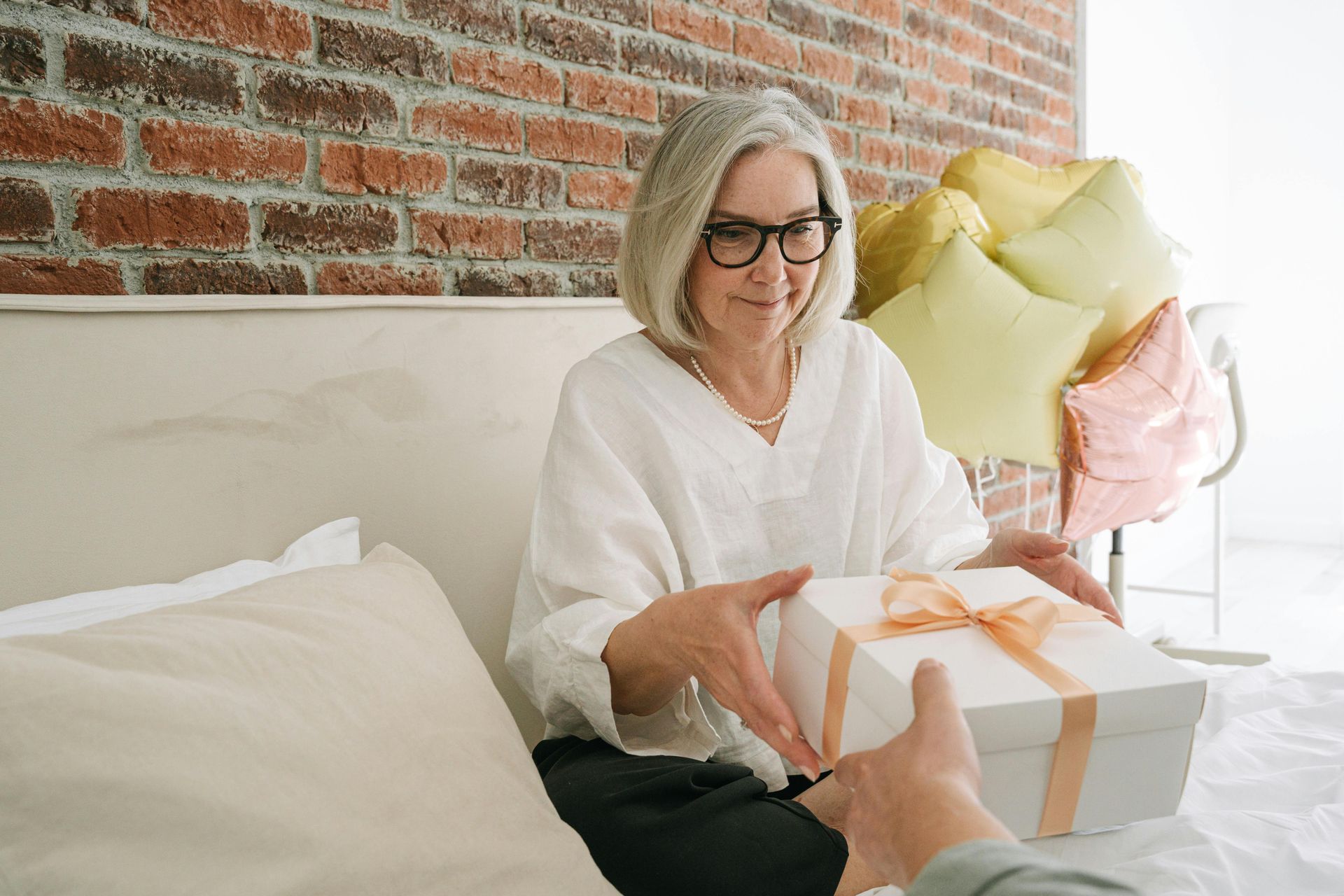 Woman with glasses gives a gift box tied with a peach bow. Pastel balloons in background.