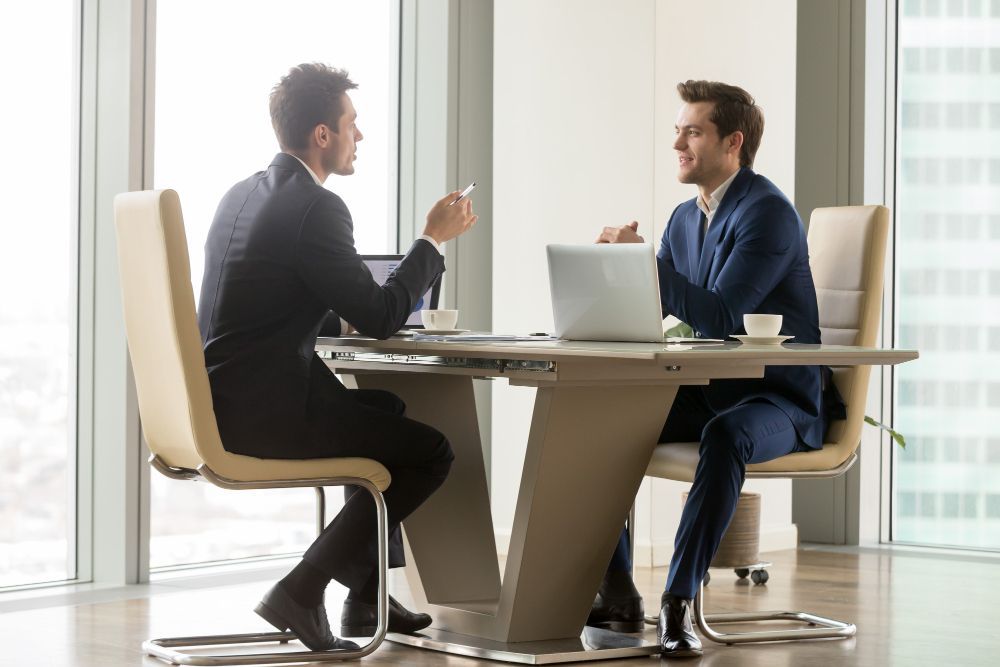 Two men are sitting at a table with laptops and having a conversation.