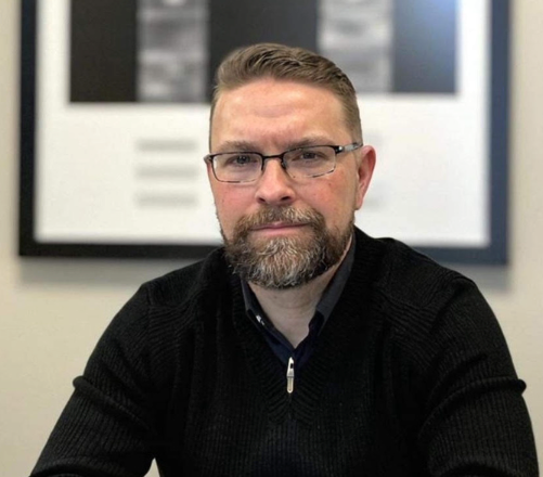 A man with a beard and glasses is sitting in front of a framed picture.