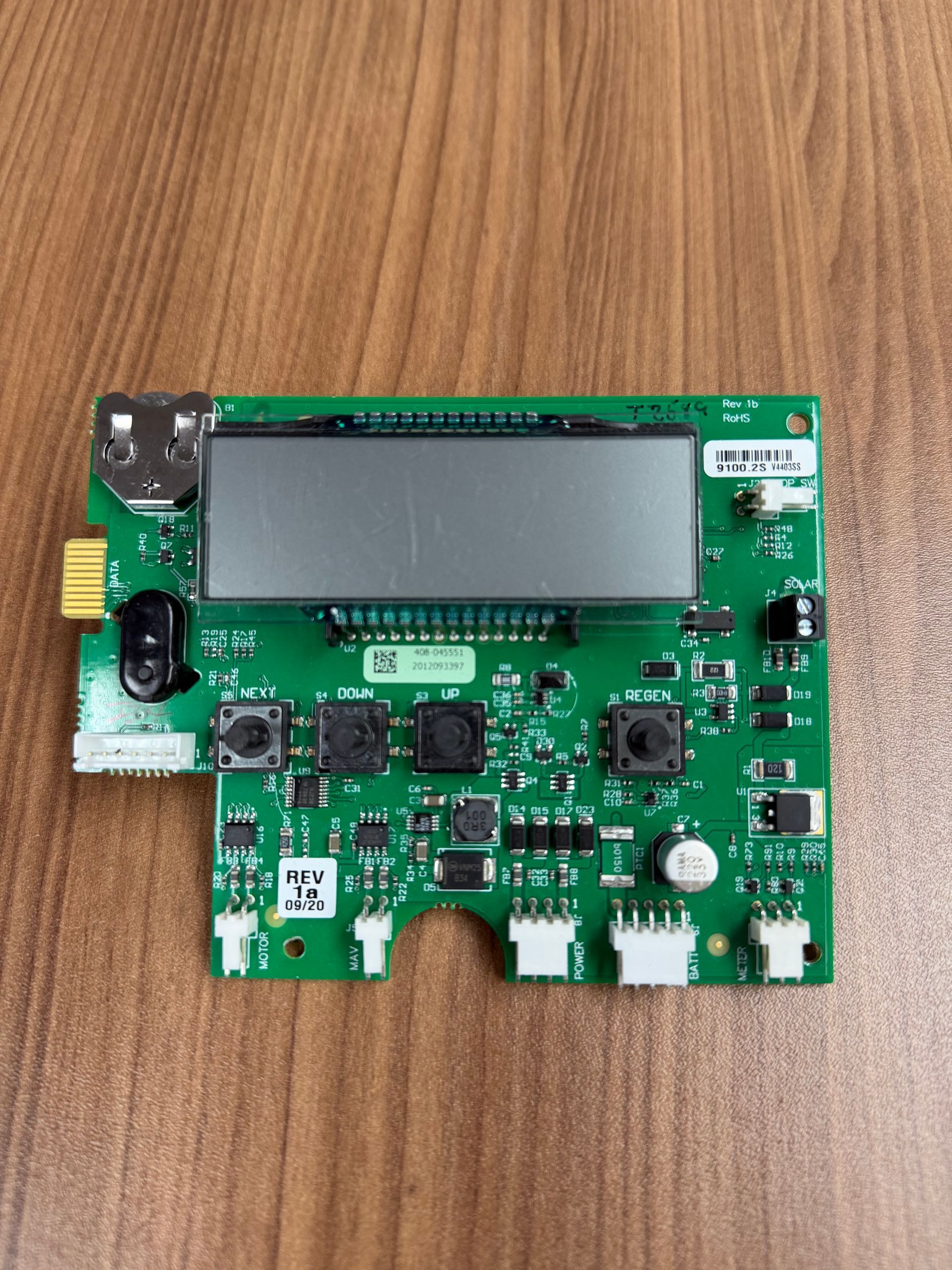 A green electronic circuit board with a central rectangular screen, various buttons, and connector ports on a wood surface.