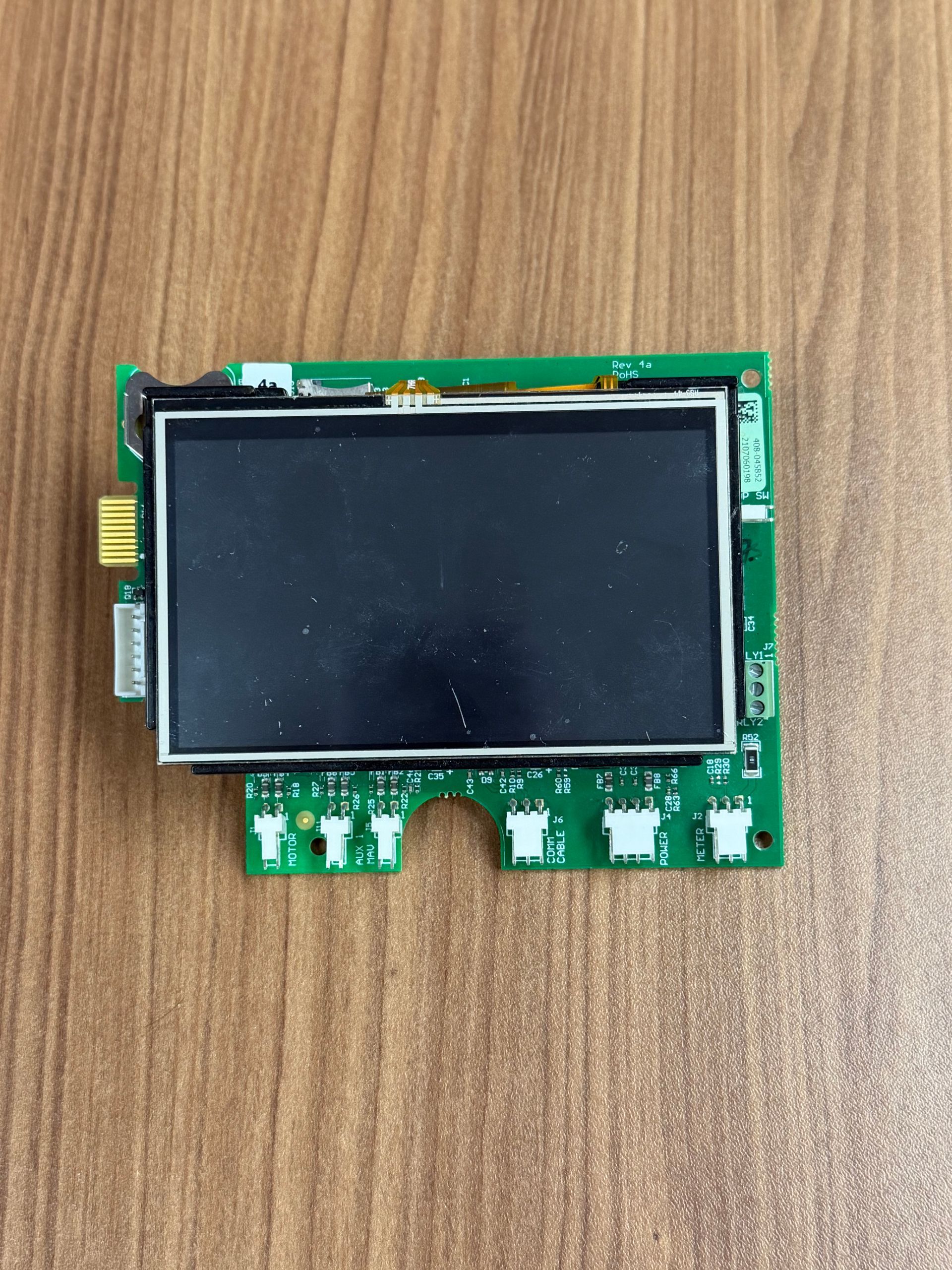 A small rectangular LCD screen mounted on a green circuit board with several white connectors, resting on a wood surface.