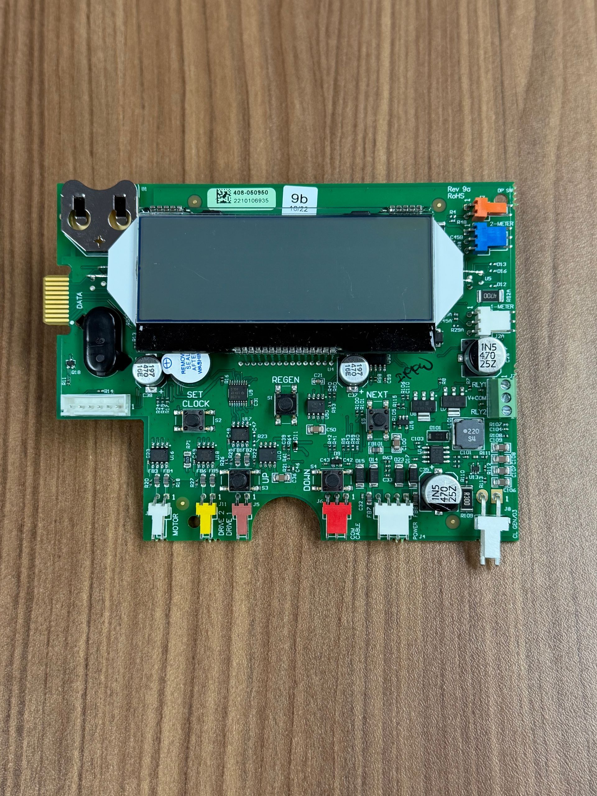 A green printed circuit board with a central display screen and various colorful connector ports on a wooden surface.