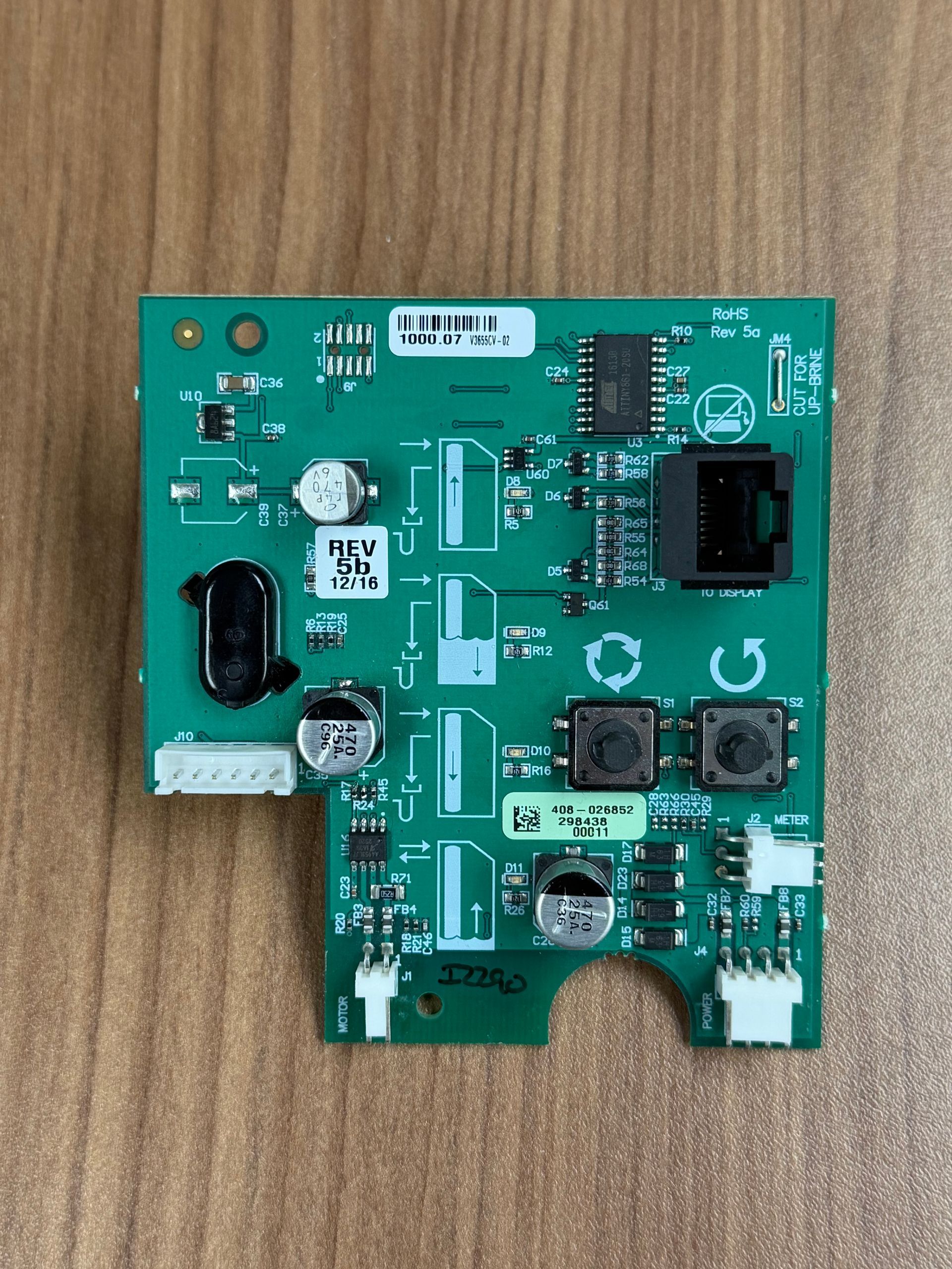 A green rectangular circuit board with various electronic components, connectors, and two black buttons on a wood surface.