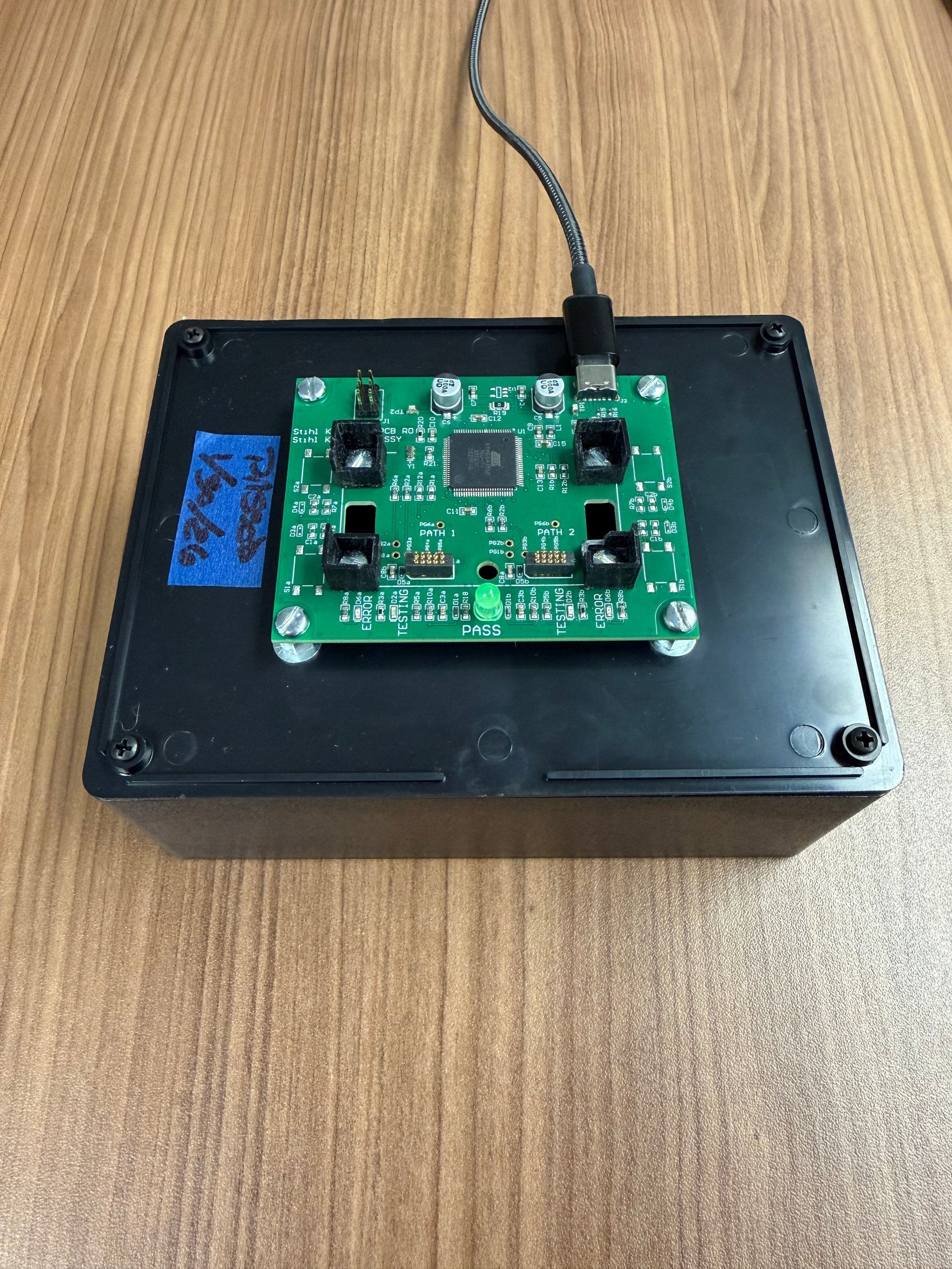 A green circuit board with a central chip and four sensors, mounted on a black box and connected by a USB cable.