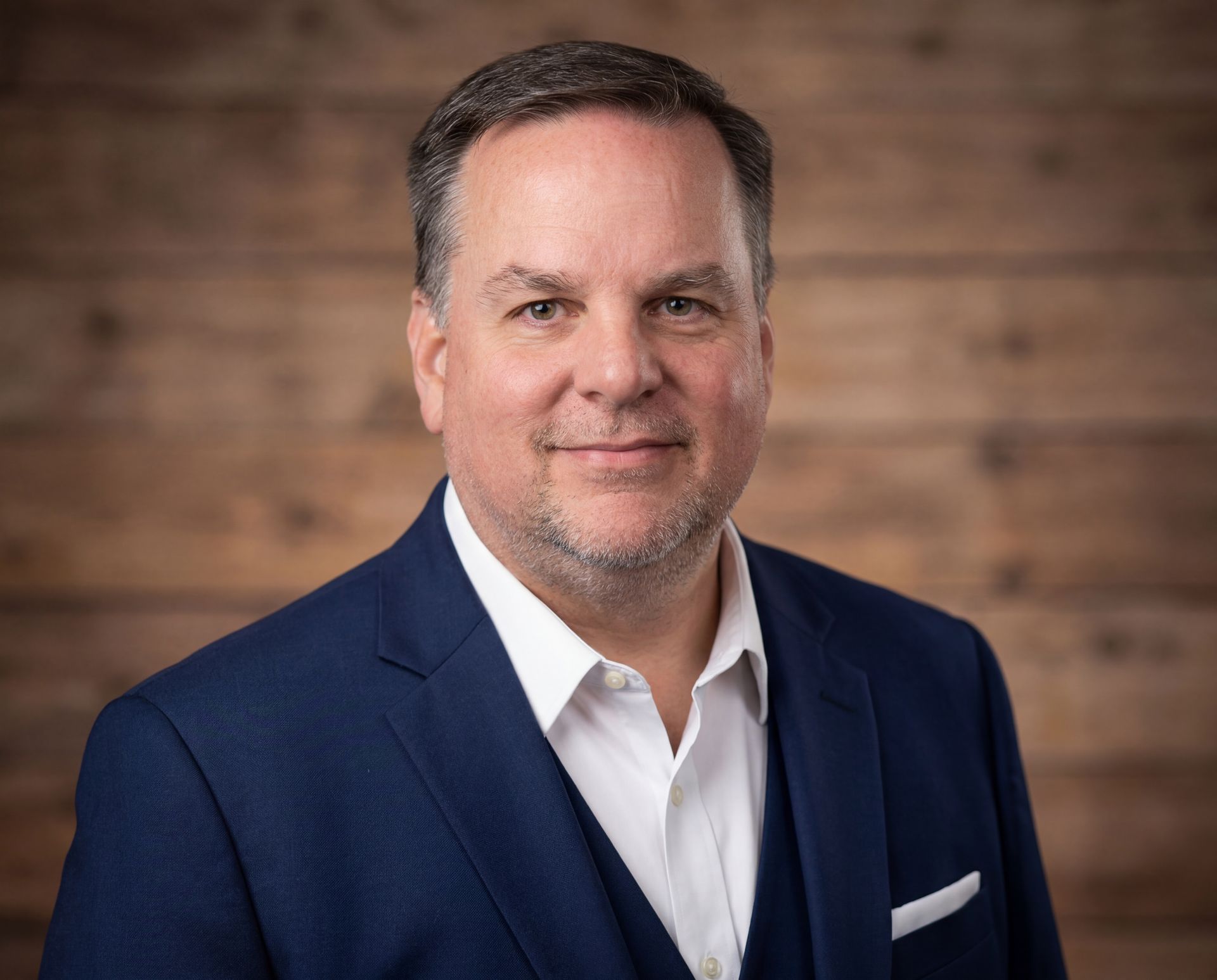 Headshot of a person in a navy suit and white shirt, posing against a wood-paneled wall.