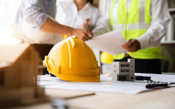Yellow hardhat on blueprint table with architects planning.