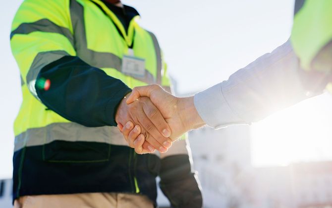 Two people in high-visibility vests shake hands, outdoors.
