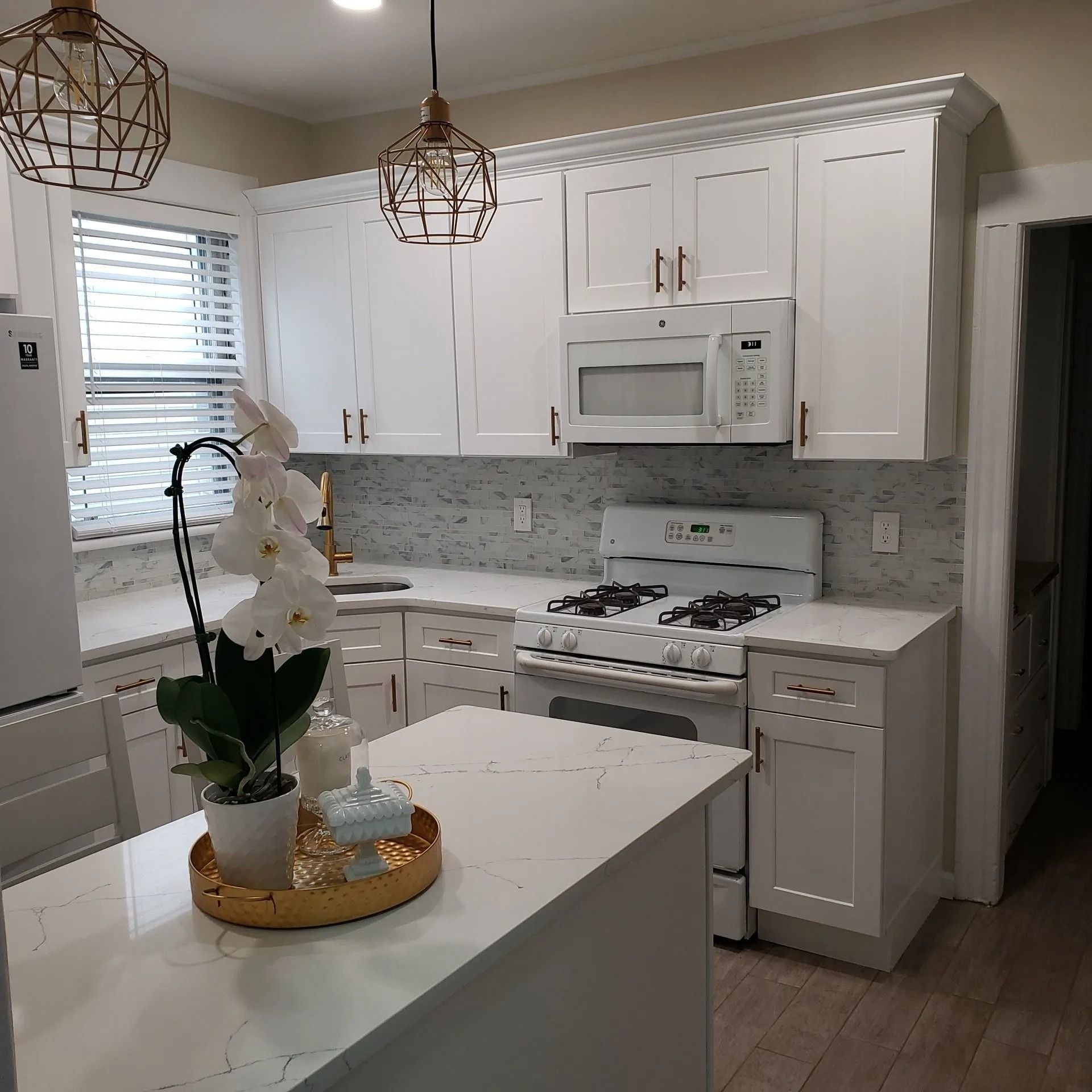 White kitchen with white cabinets, island, and countertops; marble backsplash; gold light fixtures.
