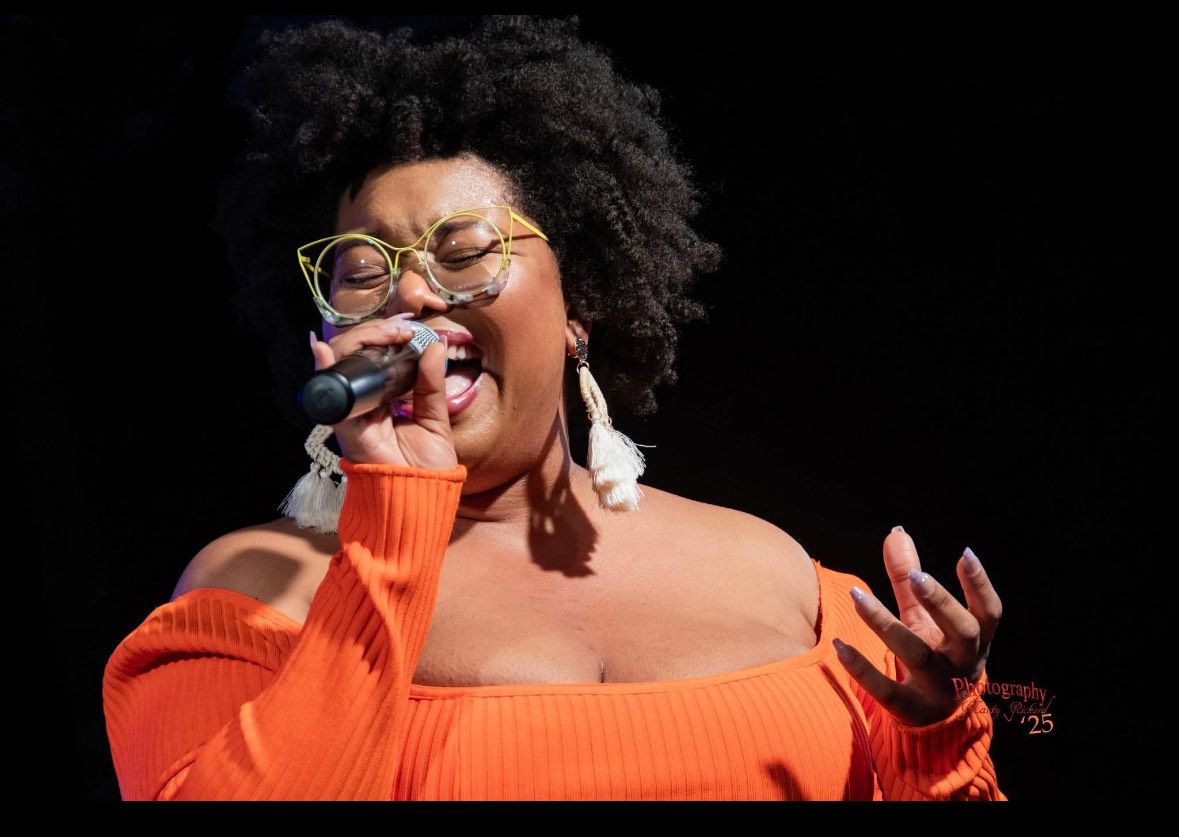 A woman wearing glasses and an orange sweater is singing into a microphone.