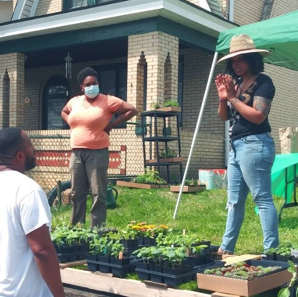 A woman wearing a hat and a mask stands in front of a house