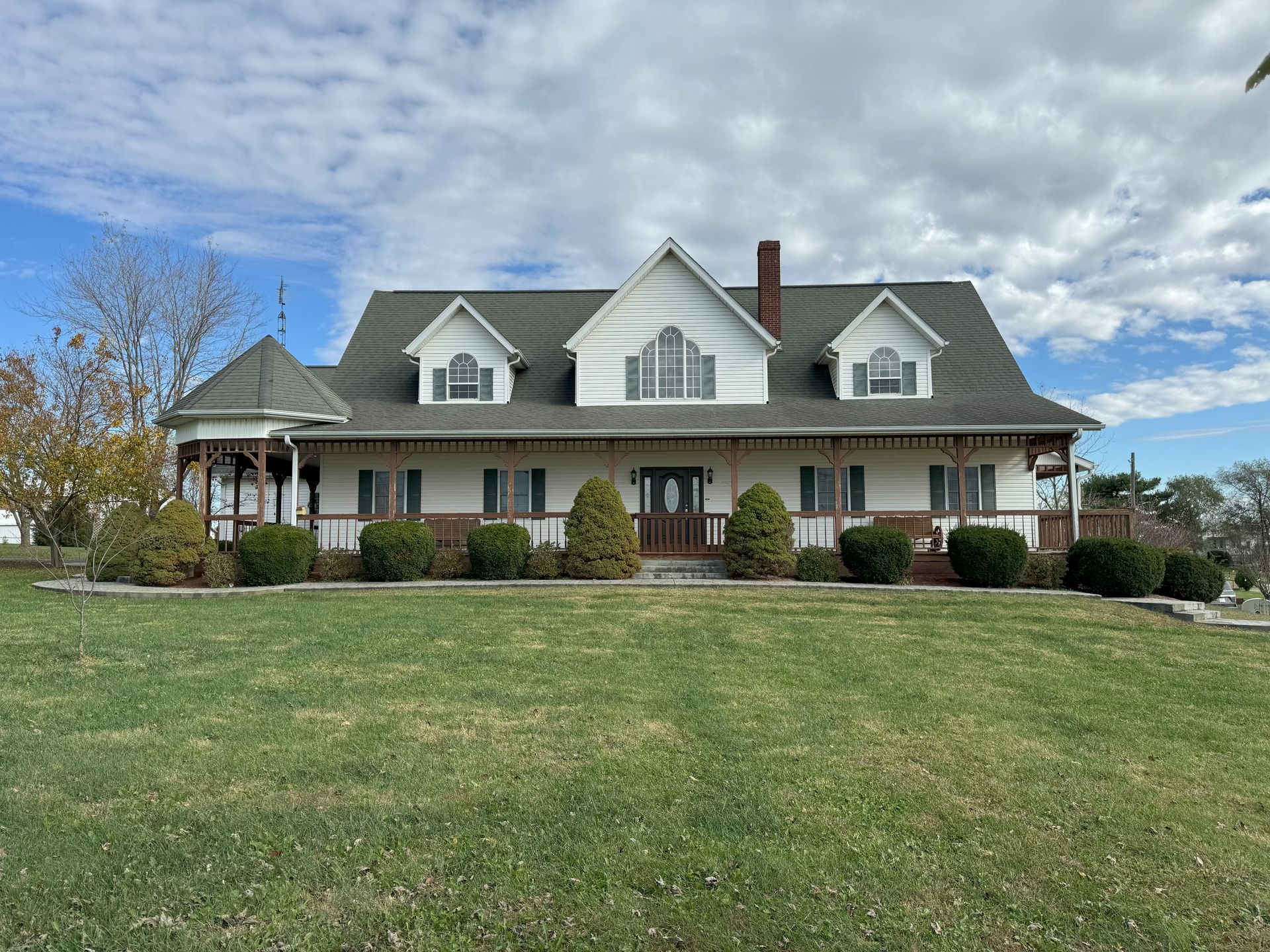 A large white house with a gray roof is sitting on top of a lush green field.