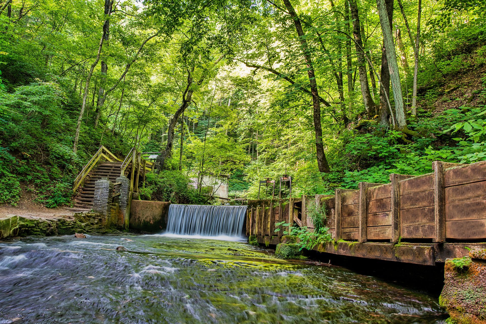 A small waterfall in the middle of a forest next to a wooden bridge.