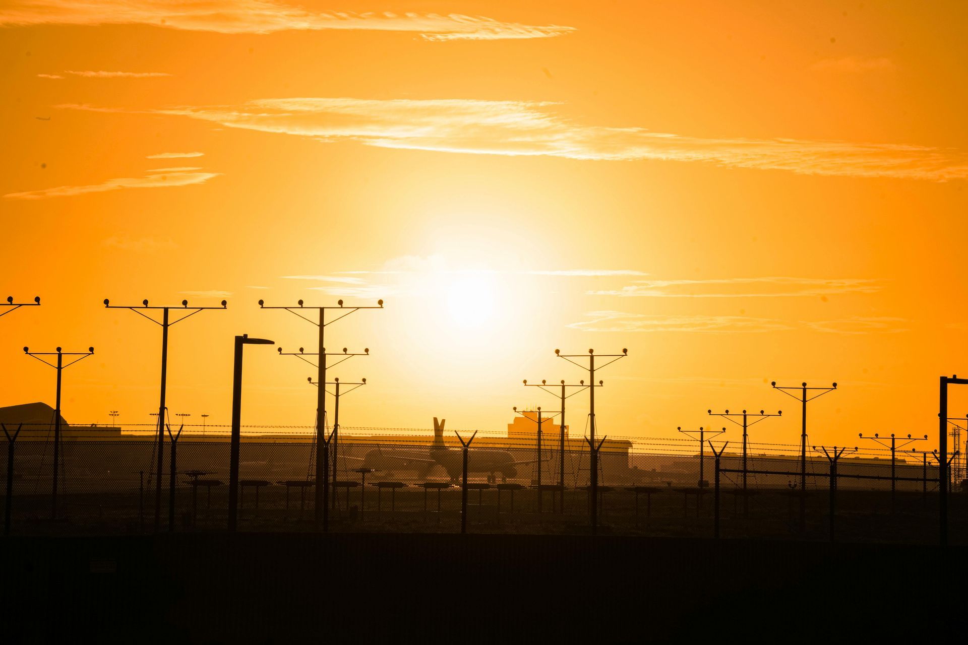 A sunset over a fenced in area with power lines and a plane in the distance.