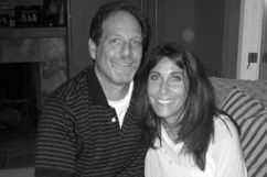 Man and woman smiling, seated indoors; man in striped shirt, woman in white sweater.