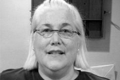 Woman with glasses and white hair smiles at the camera in a room.