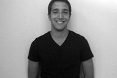 Smiling young man in a black V-neck shirt stands against a plain wall.