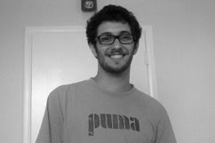 Man with glasses smiling, wearing a Puma shirt, standing in front of a white door.