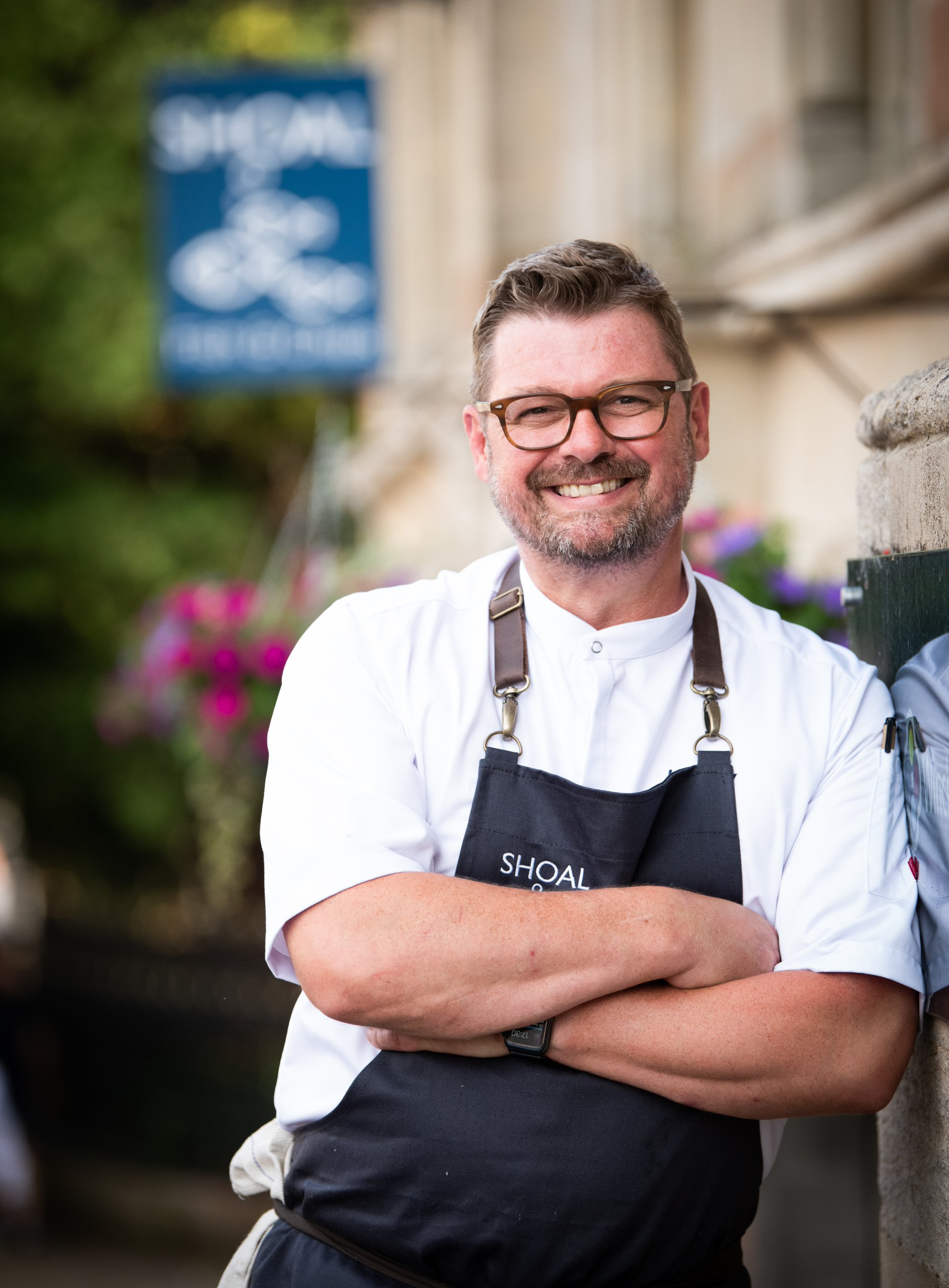 A man wearing an apron and glasses is standing with his arms crossed in front of a building.