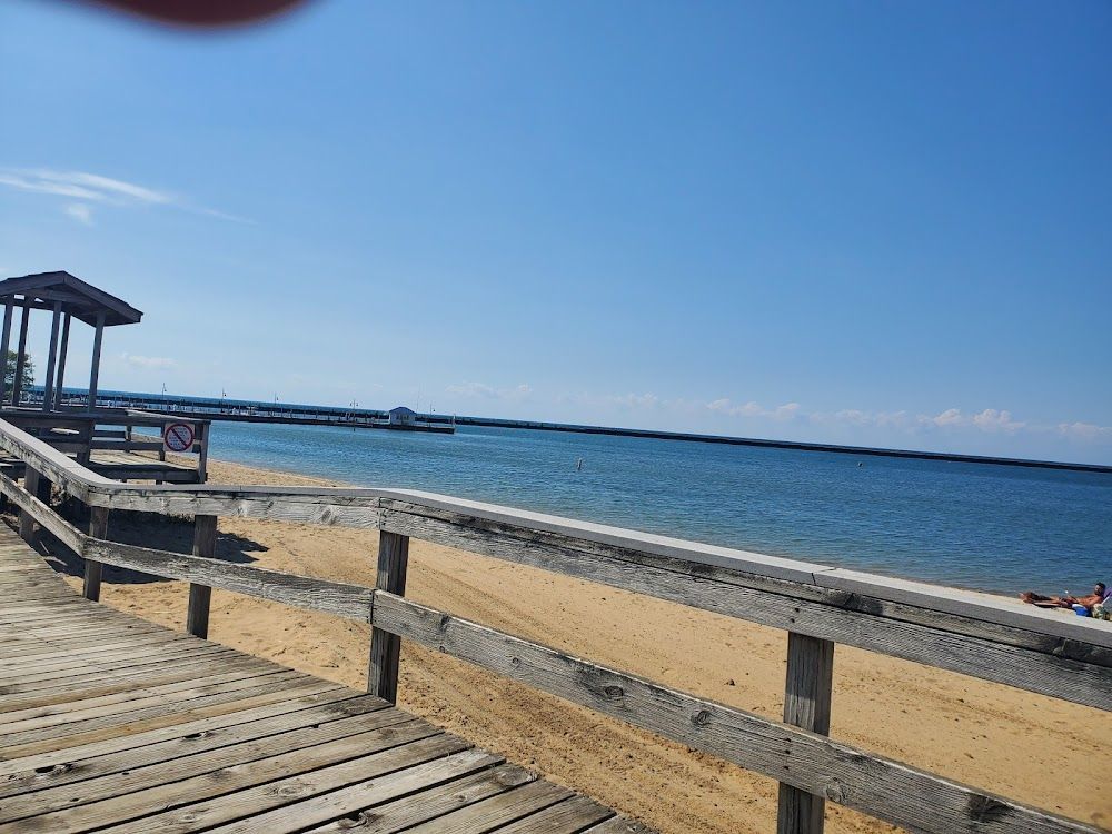 Wooden boardwalk leading to a sandy beach with blue water and sky; small shelter in view.