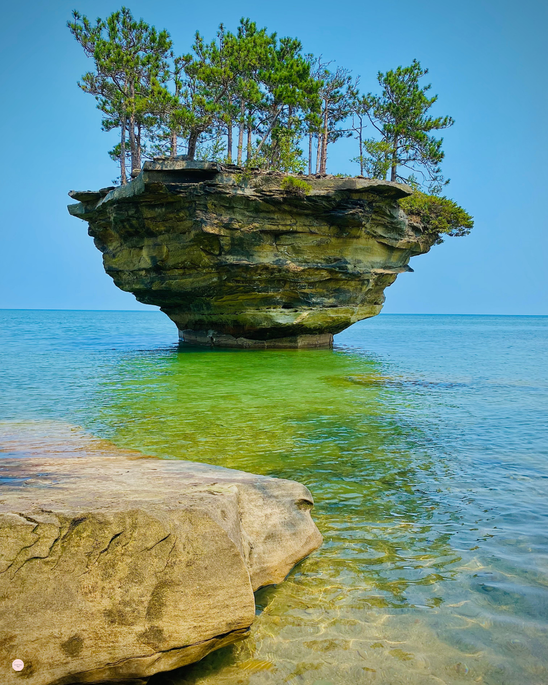 Island rock formation with trees in a lake, water is green and blue, rock is tan.