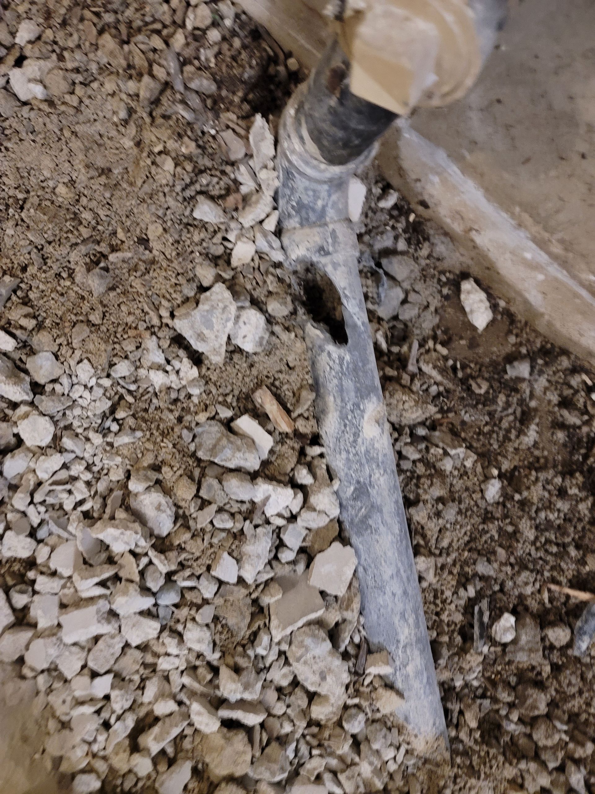 A damaged gray pipe with a large hole, embedded in loose gravel and concrete debris next to a solid surface.