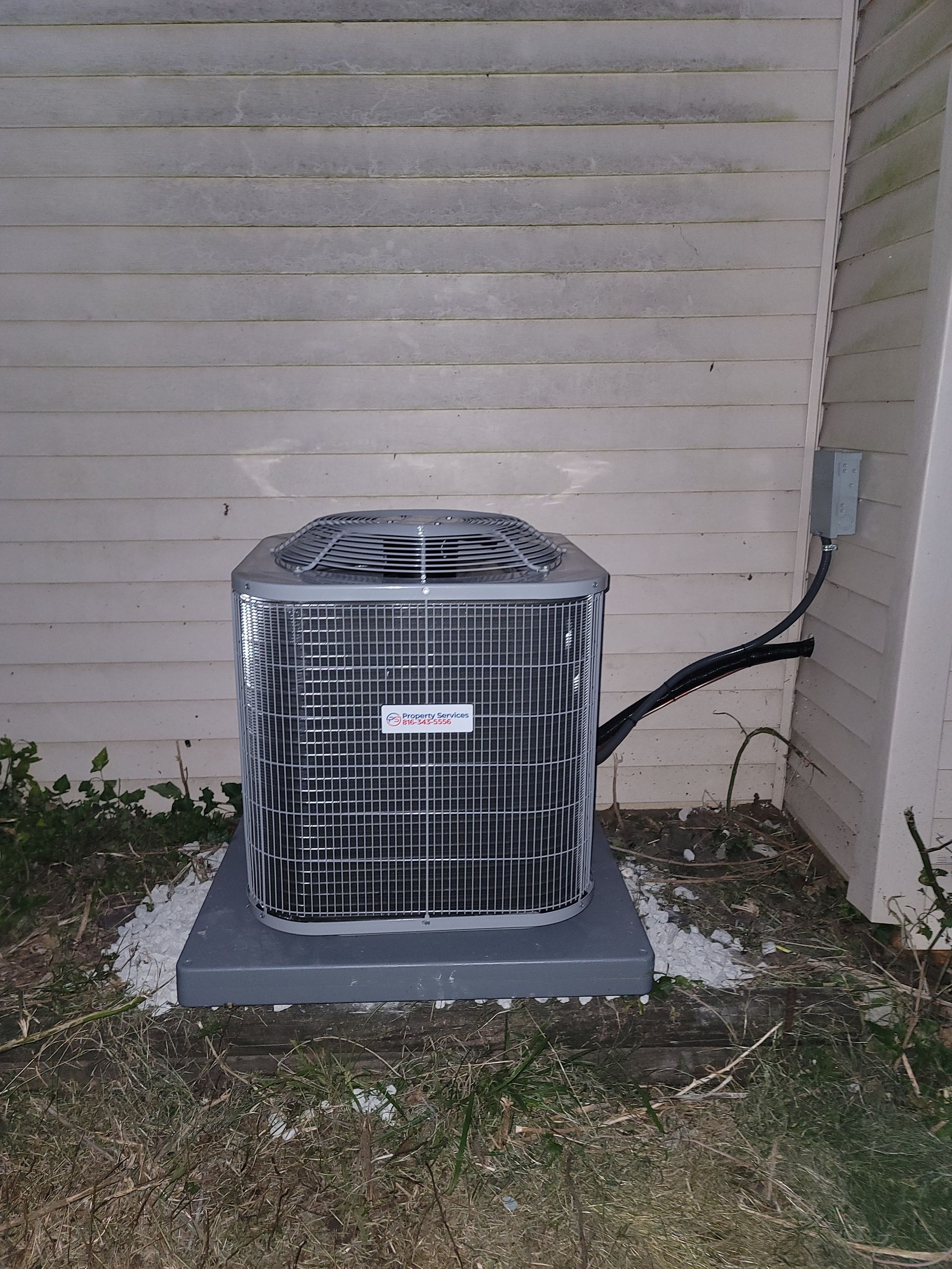An outdoor air conditioning unit sits on a gray concrete slab against a light-colored wooden siding wall at night.