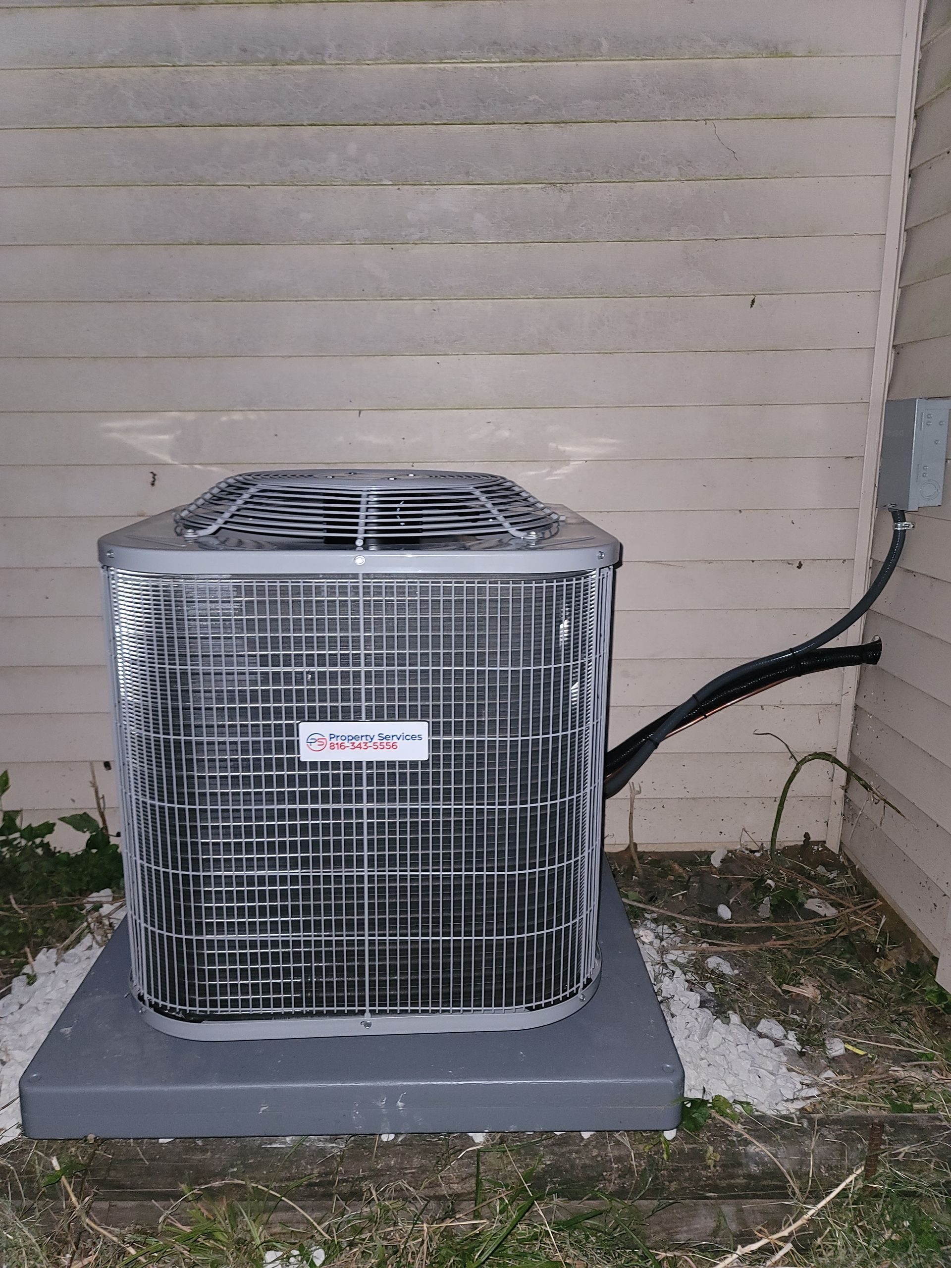 An outdoor central air conditioning unit sits on a concrete pad against a house with light-colored siding.