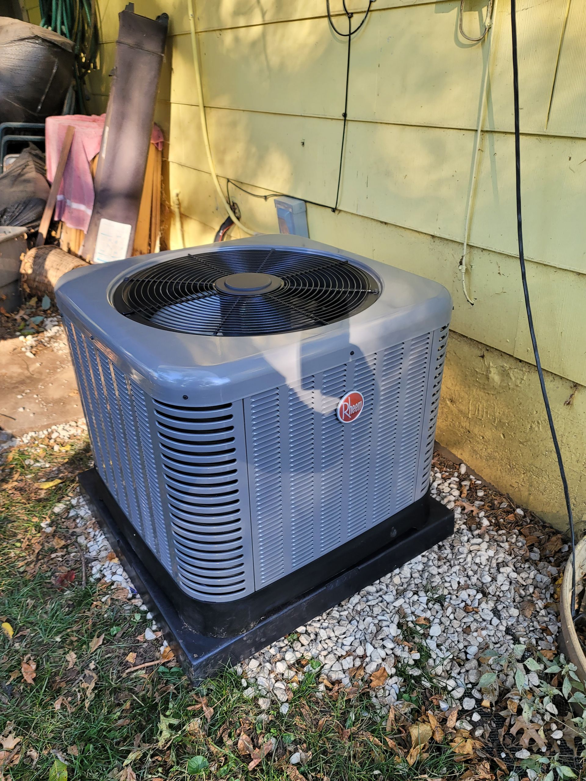 A grey Rheem outdoor air conditioning unit sits on a gravel base next to a yellow wall.