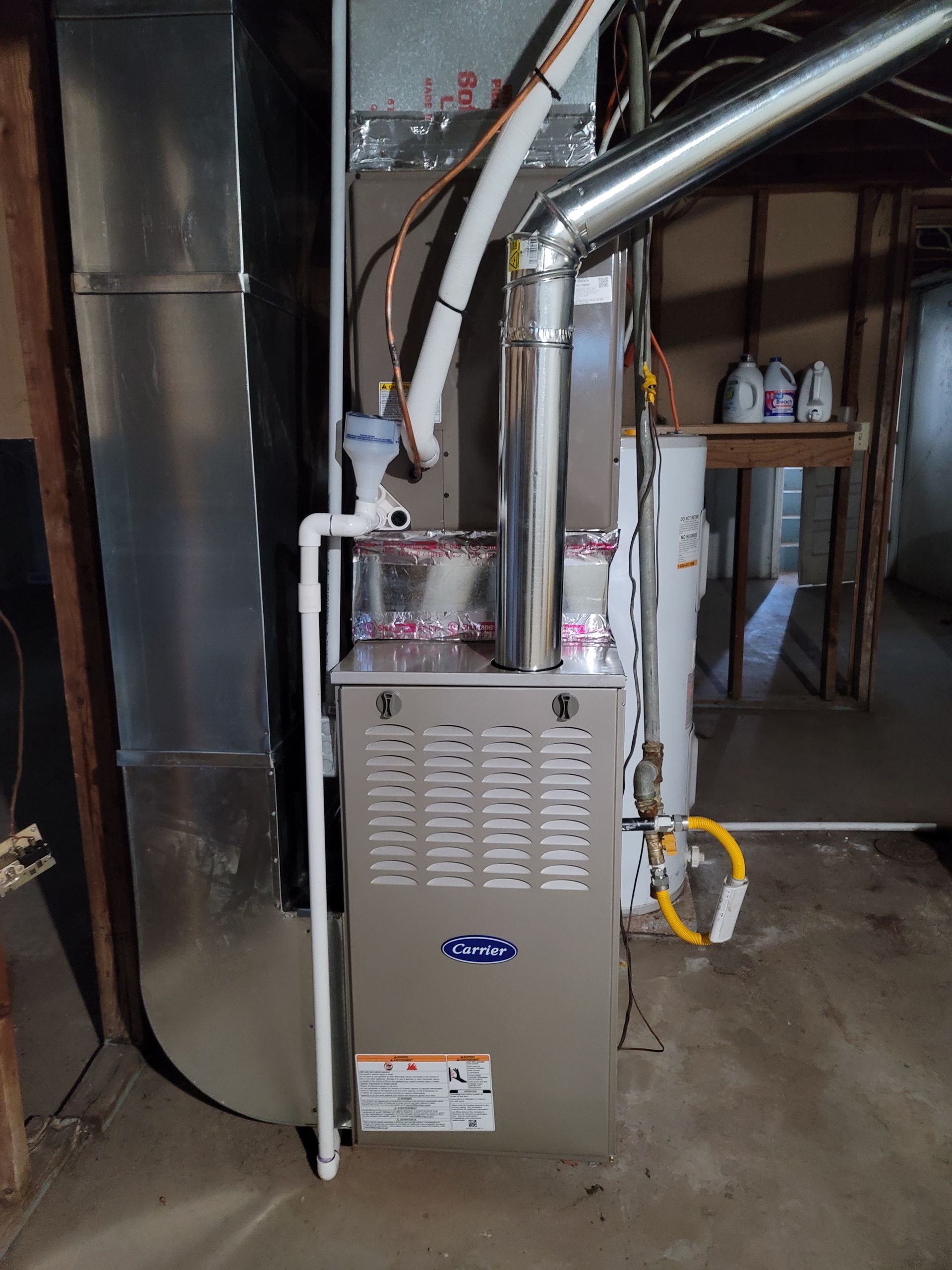 A silver residential furnace with metal ductwork stands in a basement next to a white PVC drain pipe and gas line.