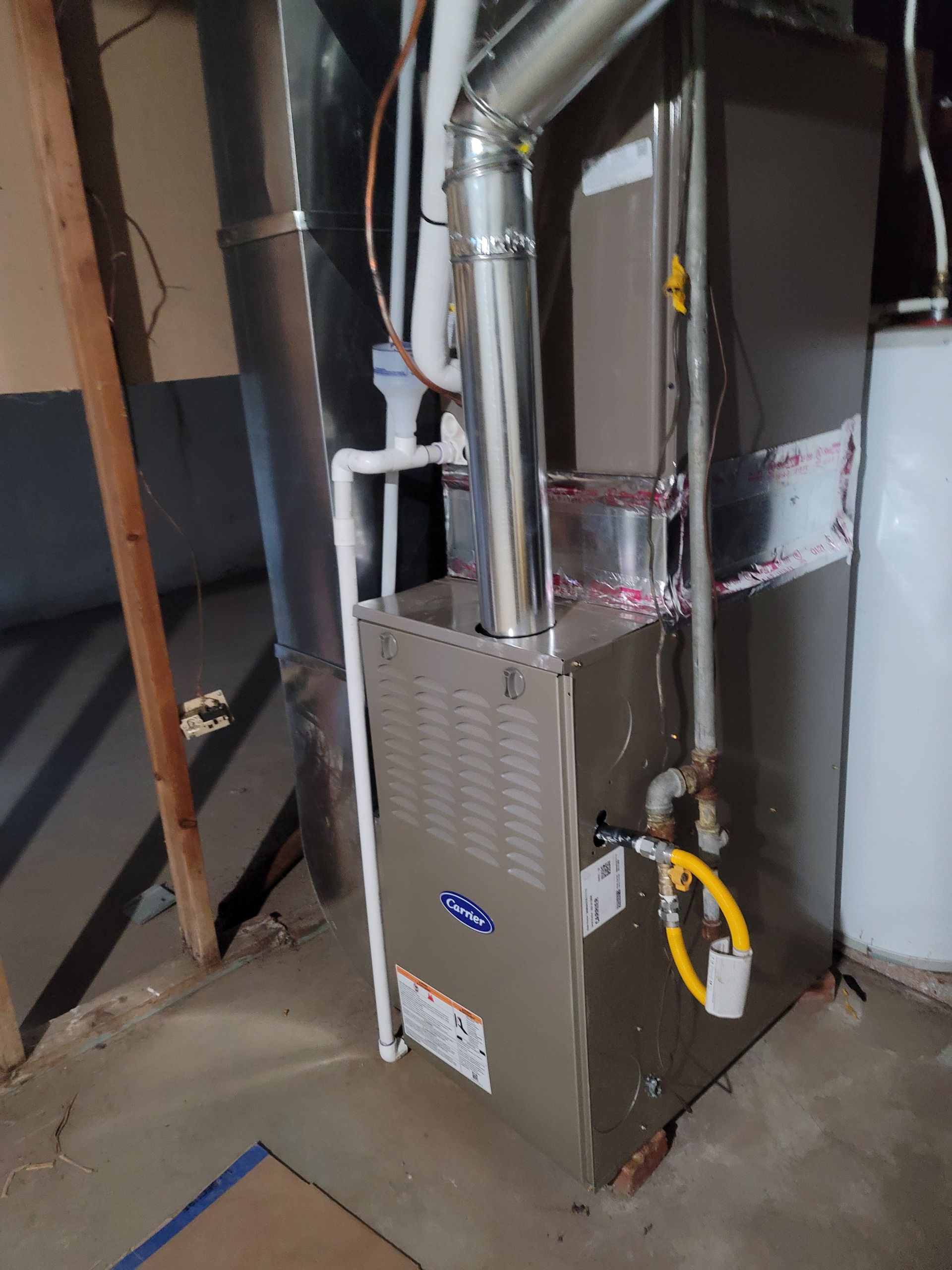 A gas furnace installed in a residential basement with visible silver ducting, gas piping, and white PVC exhaust pipes.