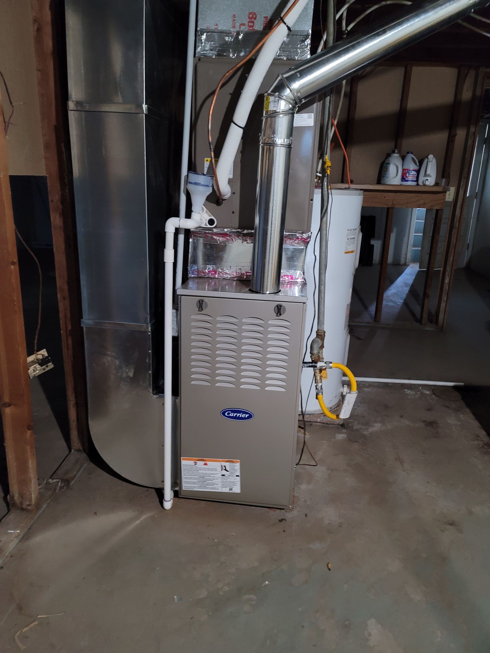 A gray furnace with connected silver ductwork and a white PVC drain pipe stands in a basement.