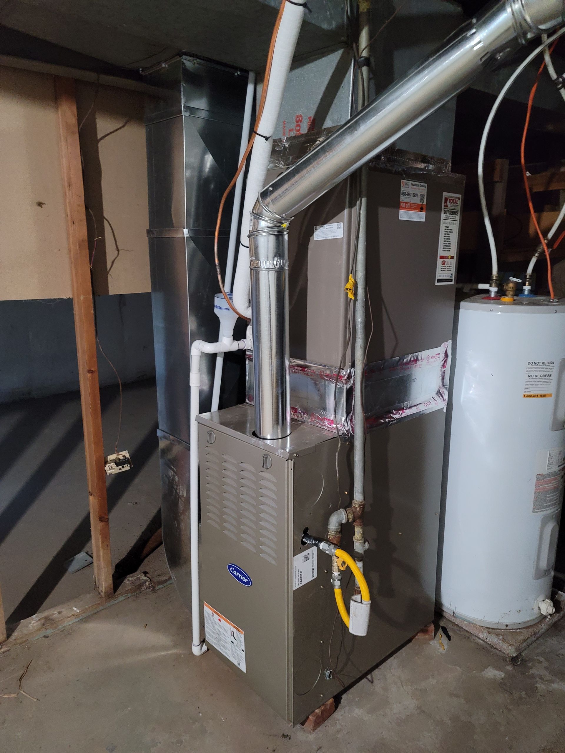 A grey furnace with silver vent pipes stands in a basement next to a white water heater and metal ductwork.