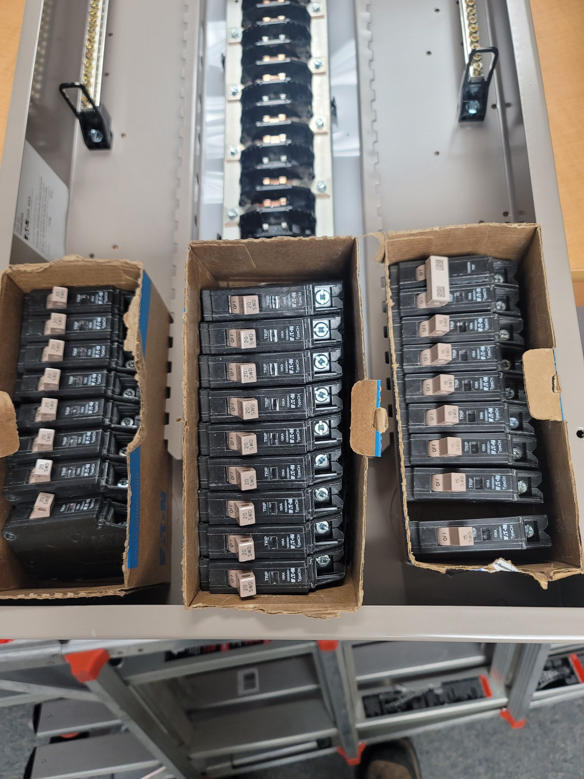 An empty electrical panel sits above three boxes containing several new circuit breakers on a workbench.