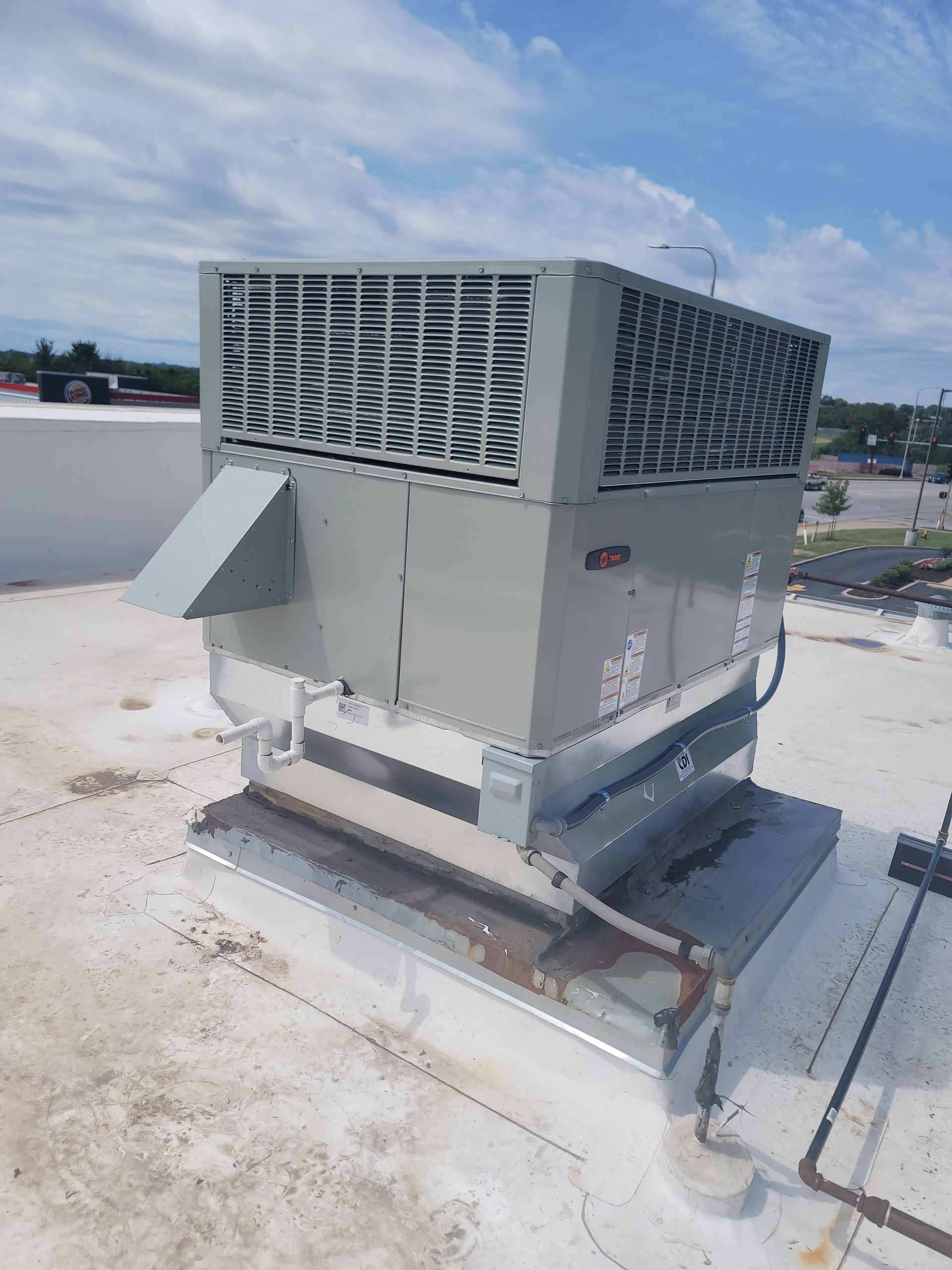 A gray industrial rooftop HVAC unit with a slanted exhaust vent sits on a flat, white roof under a bright blue sky.