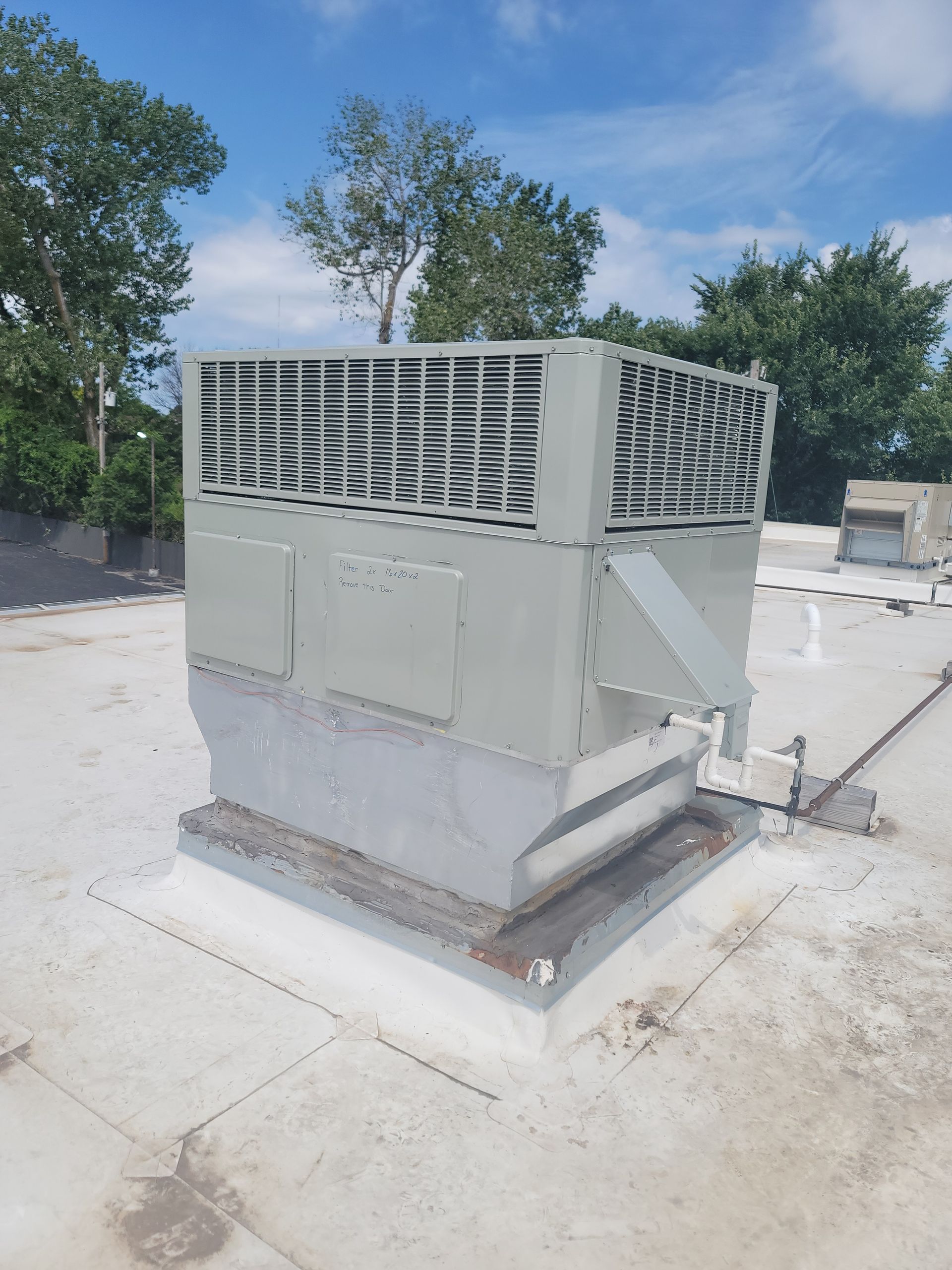 A pale green rooftop ventilation fan unit with metal louvers, set against a blue sky with trees in the background.