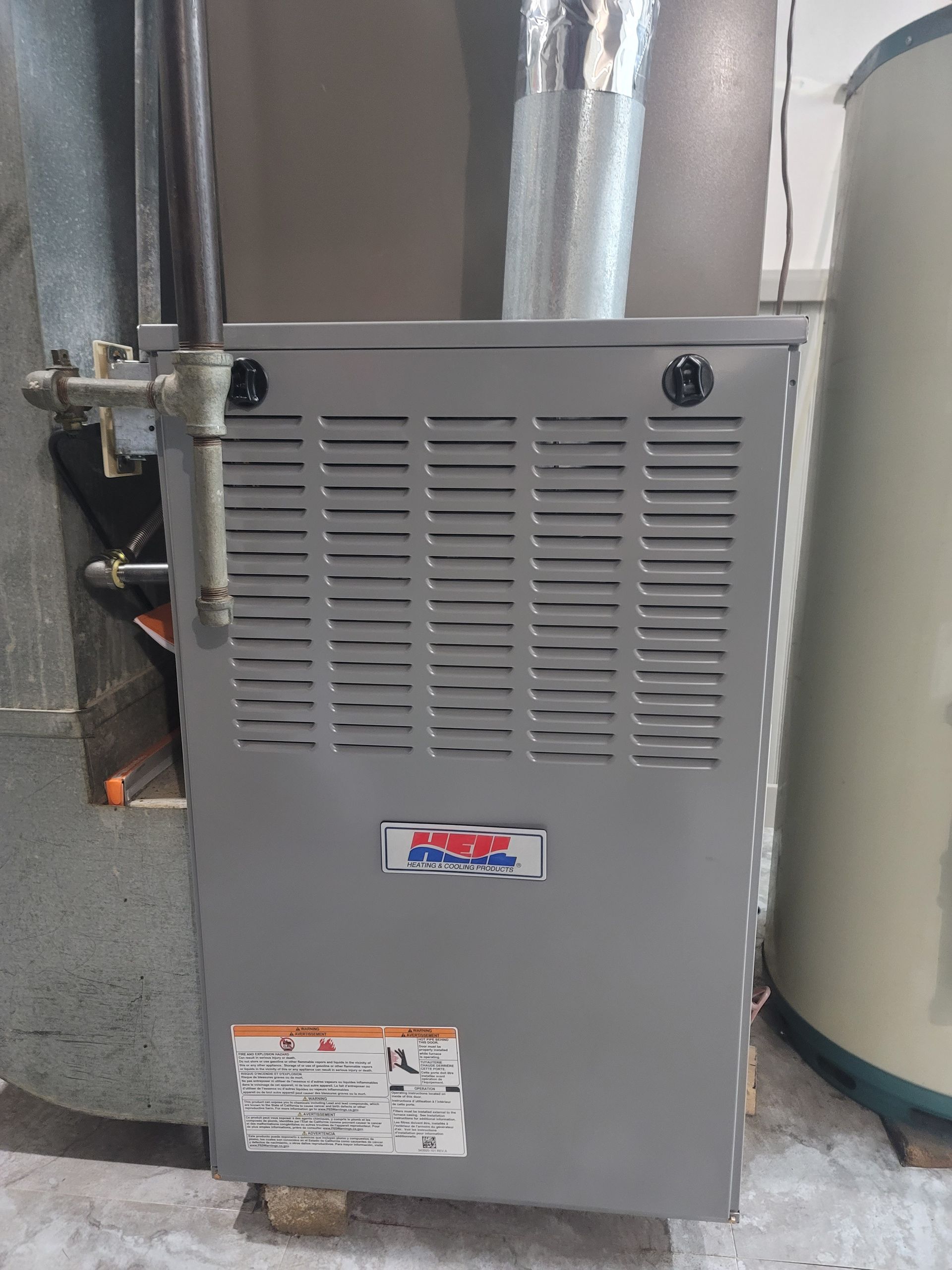 A gray gas furnace unit sits in a residential basement next to metal ductwork and a water heater.
