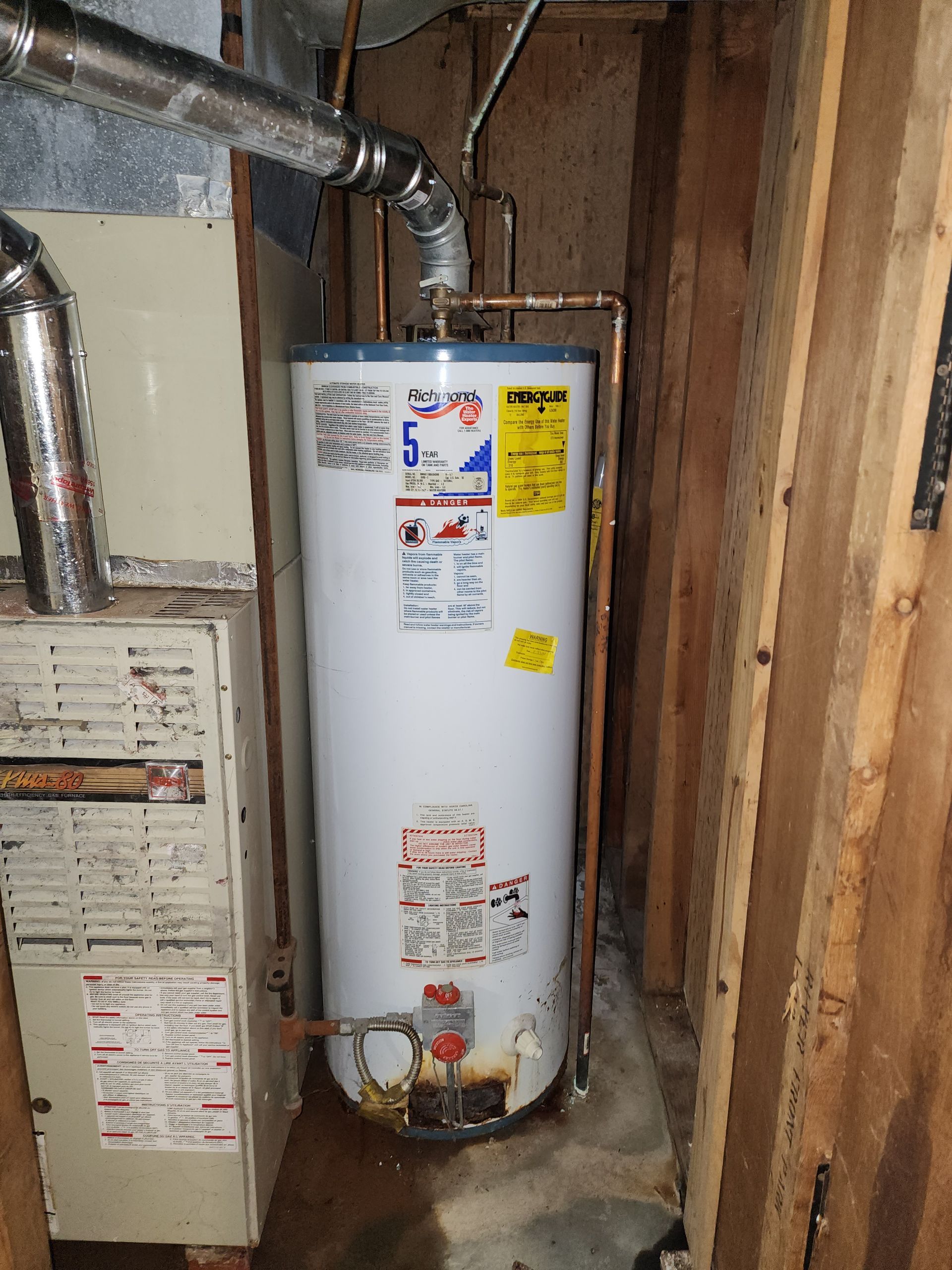 A white residential water heater installed in a utility closet next to a furnace, showing signs of rust at the base.