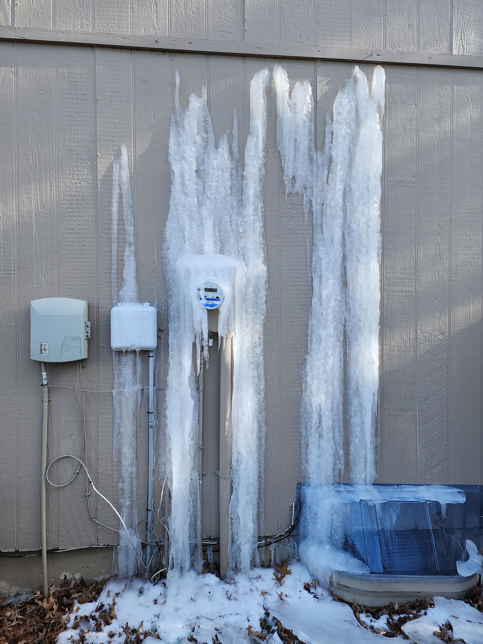 Thick ice coats the side of a building, covering utility boxes and an outdoor electrical conduit.
