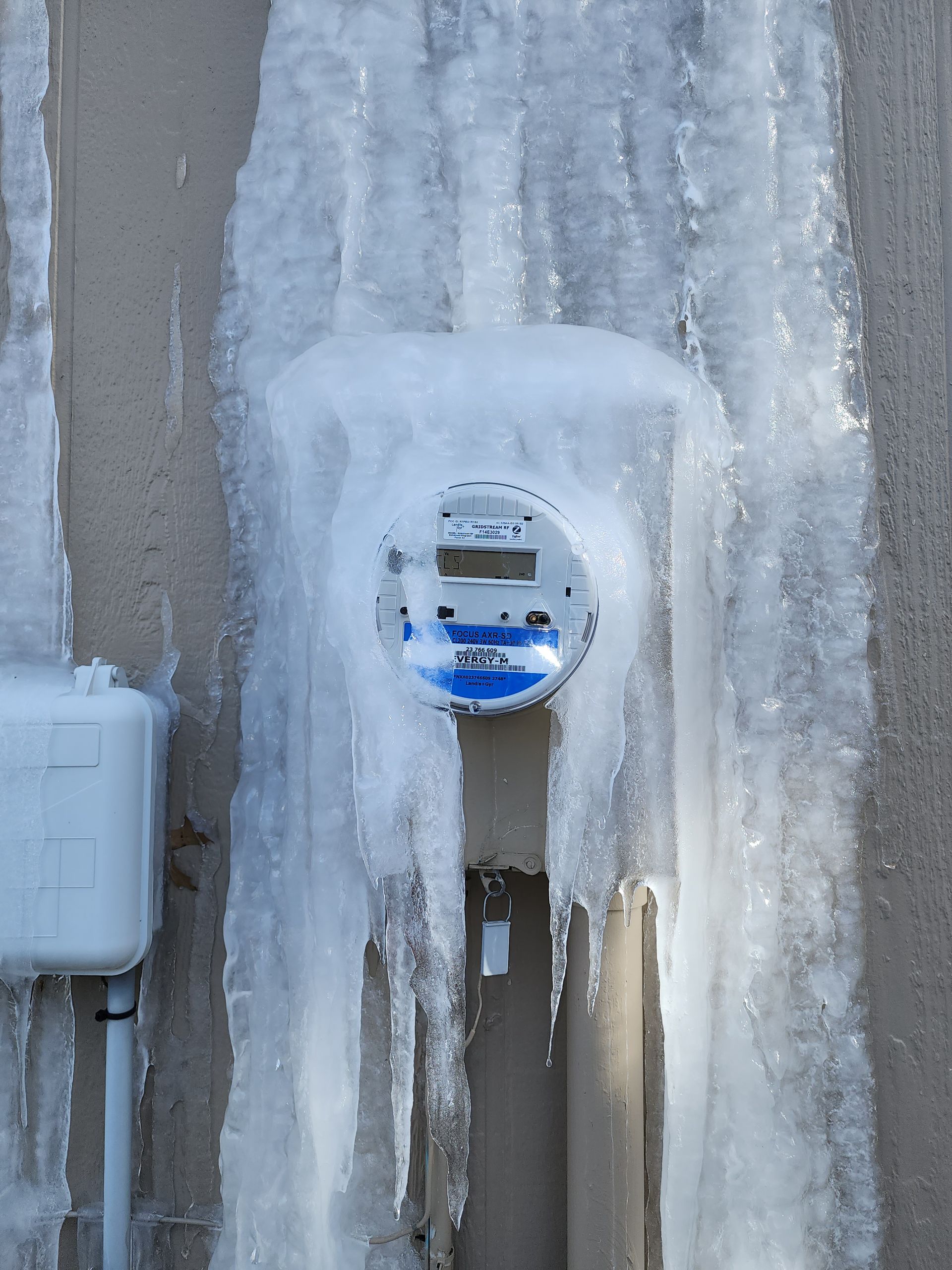 An electrical meter mounted on a beige wall is encased in a thick, heavy layer of ice with large icicles hanging down.