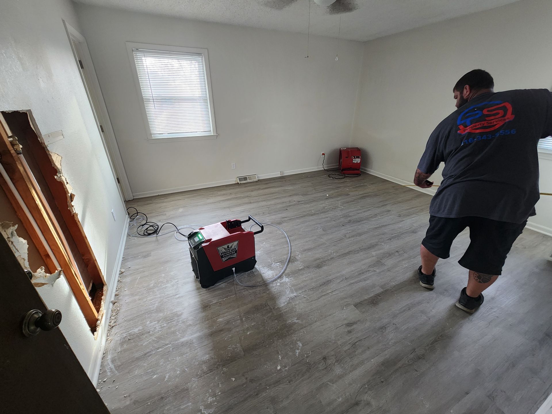 A technician in a gray shirt uses a red air mover to dry the floor of a room with light wood-style flooring.