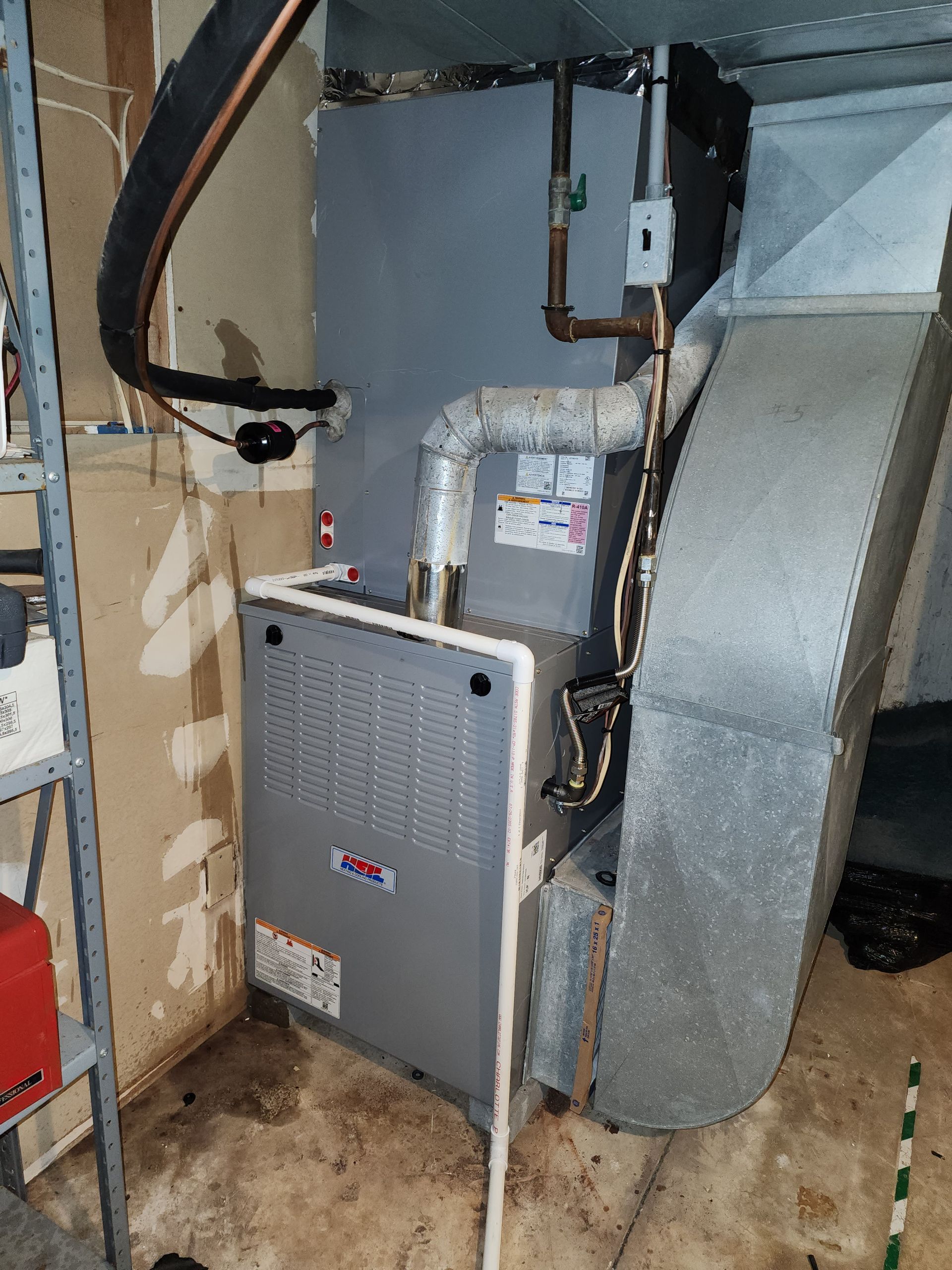 A gray indoor HVAC furnace unit installed in a utility space, connected to metal ductwork and copper piping.