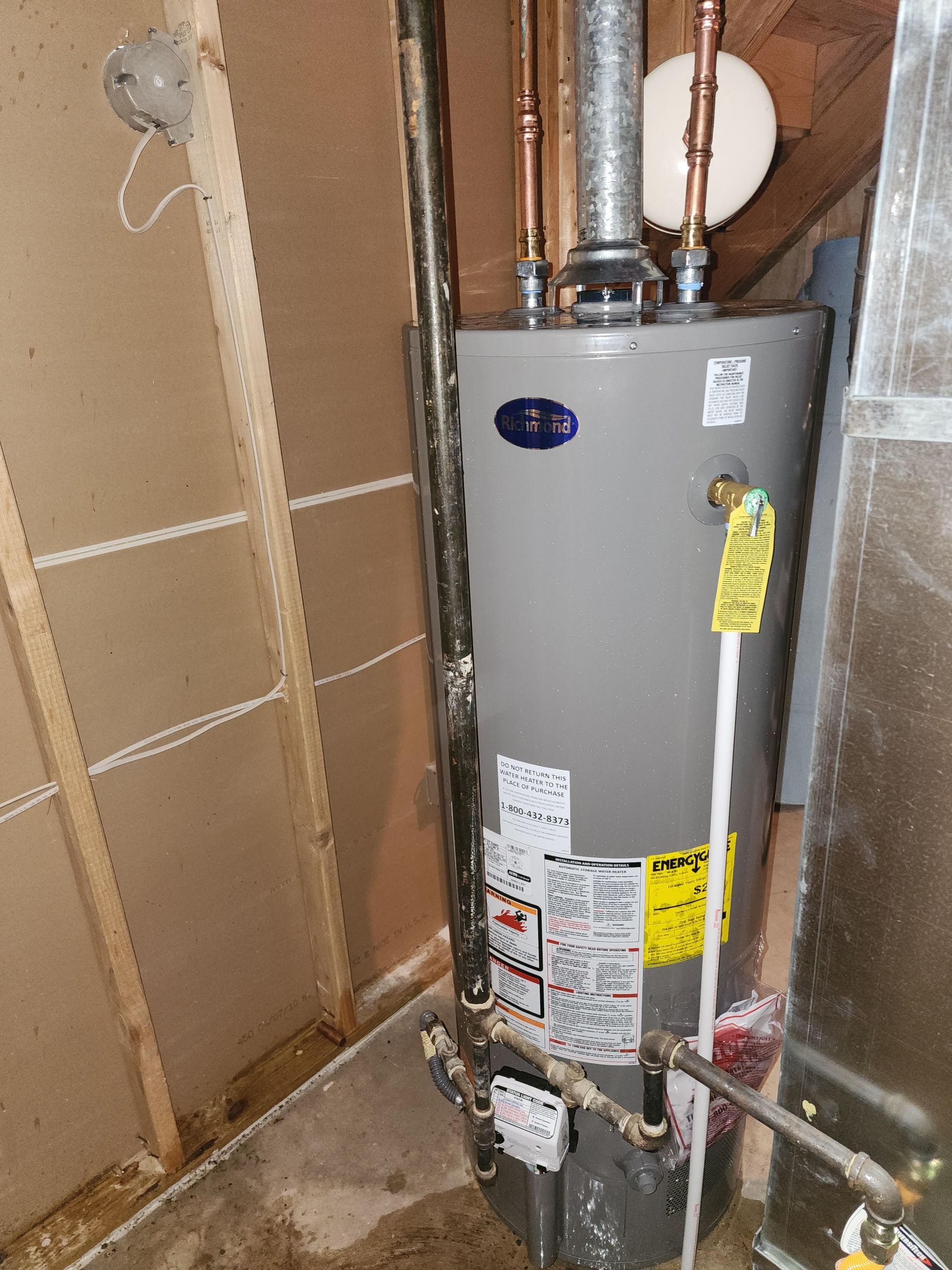 A grey gas water heater in a basement utility area with copper piping and a white discharge tube.