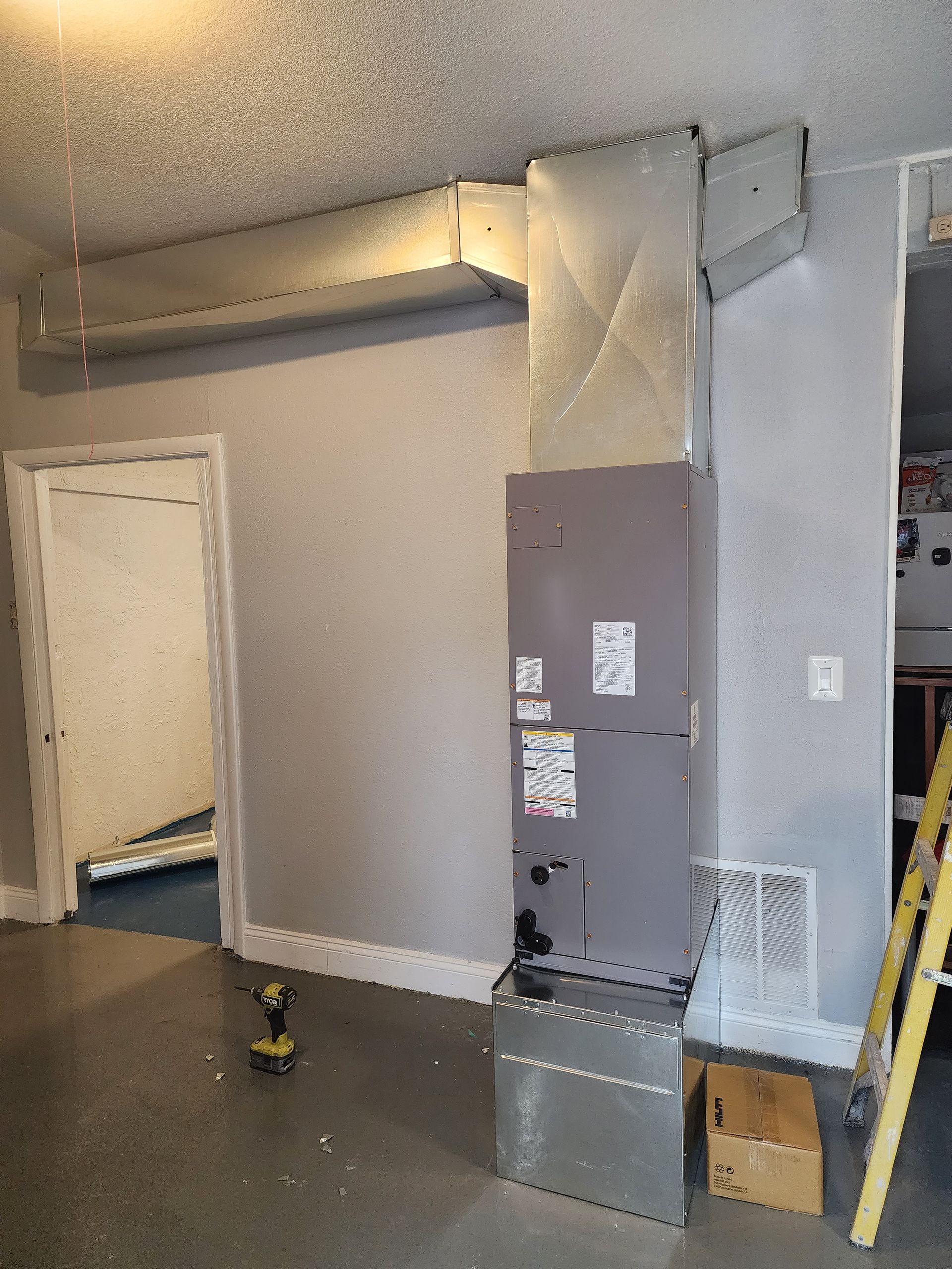 A tall, grey HVAC unit stands in a room with a white metal duct system overhead and a floor vent on the wall.