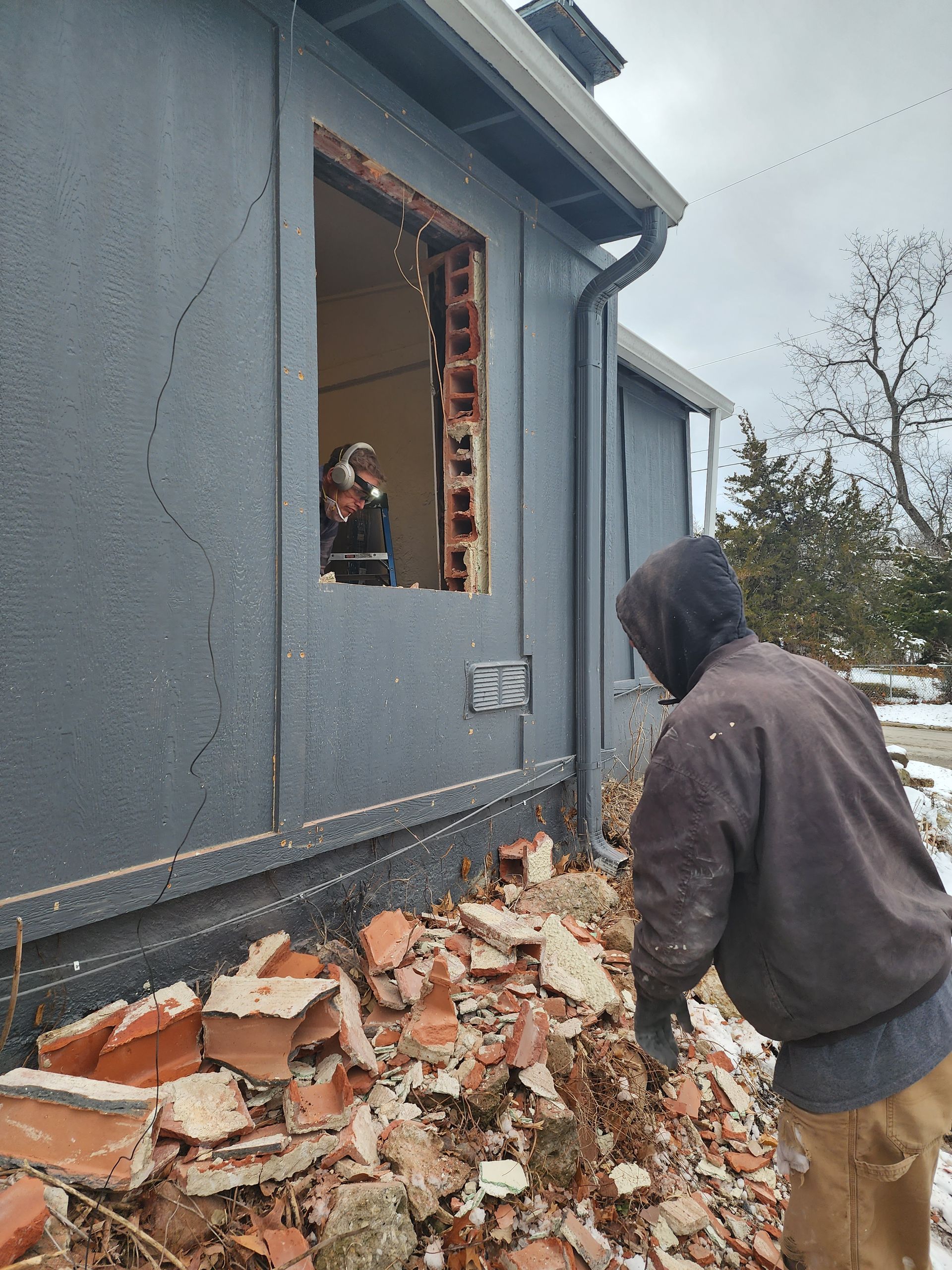 A worker in a brown jacket stands outside a house with a hole in the exterior wall, surrounded by broken brick debris.
