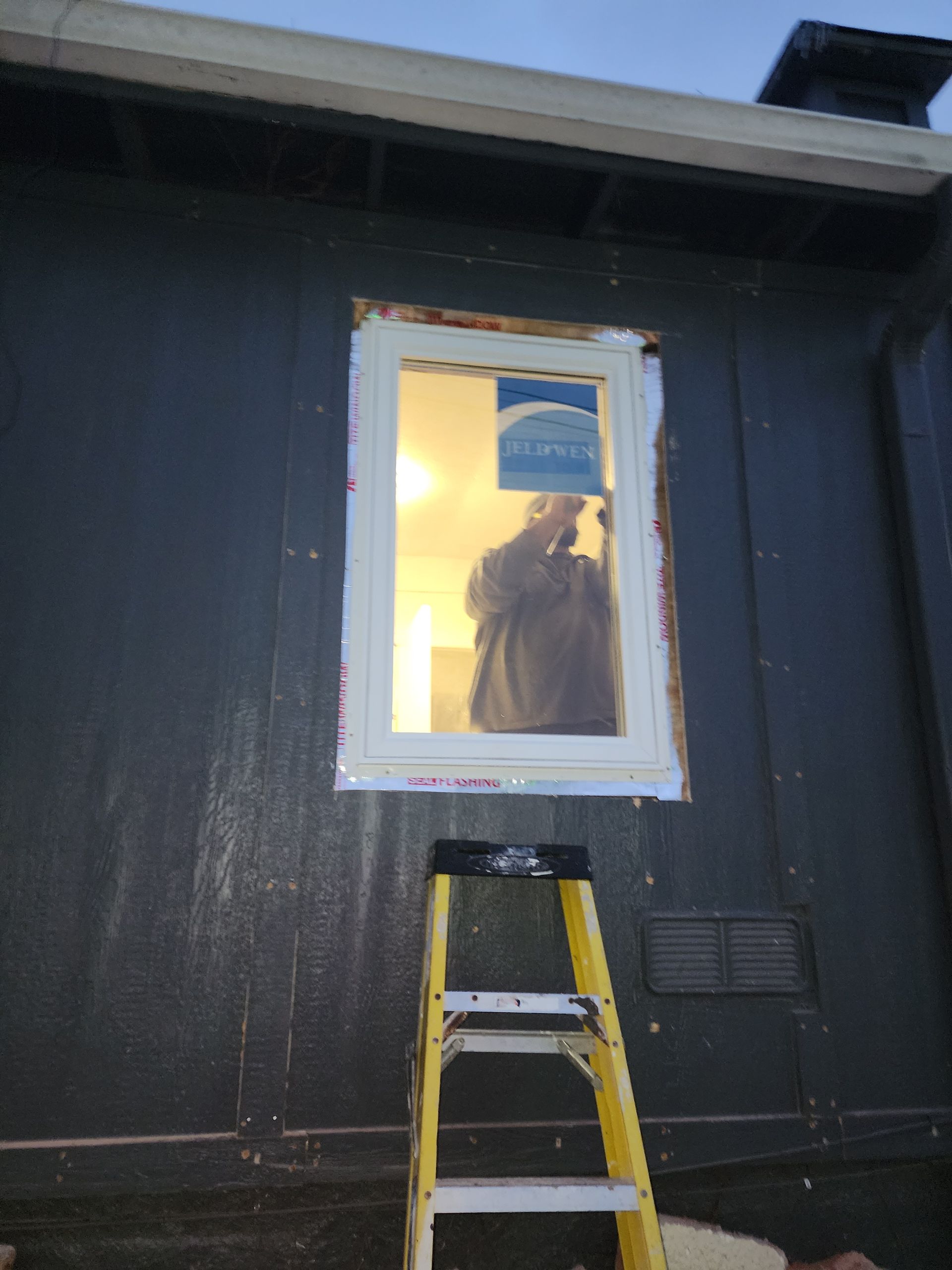 A person stands inside a new window frame installed on the exterior of a dark gray house, with a yellow ladder below.