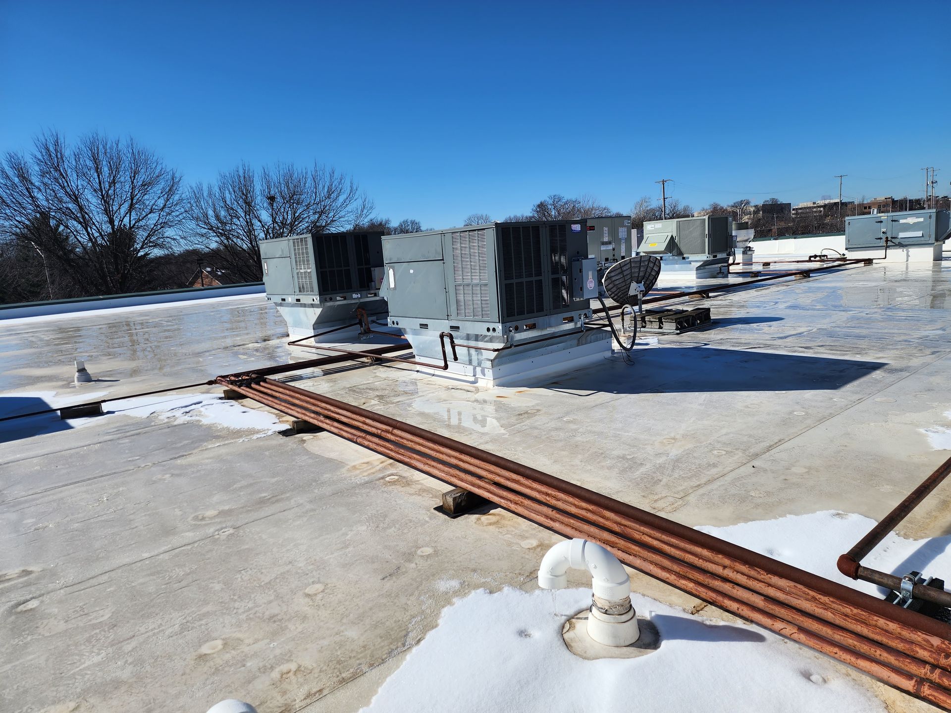 Two HVAC units sit on a snowy, flat commercial roof with several pipes running across the surface under a clear sky.